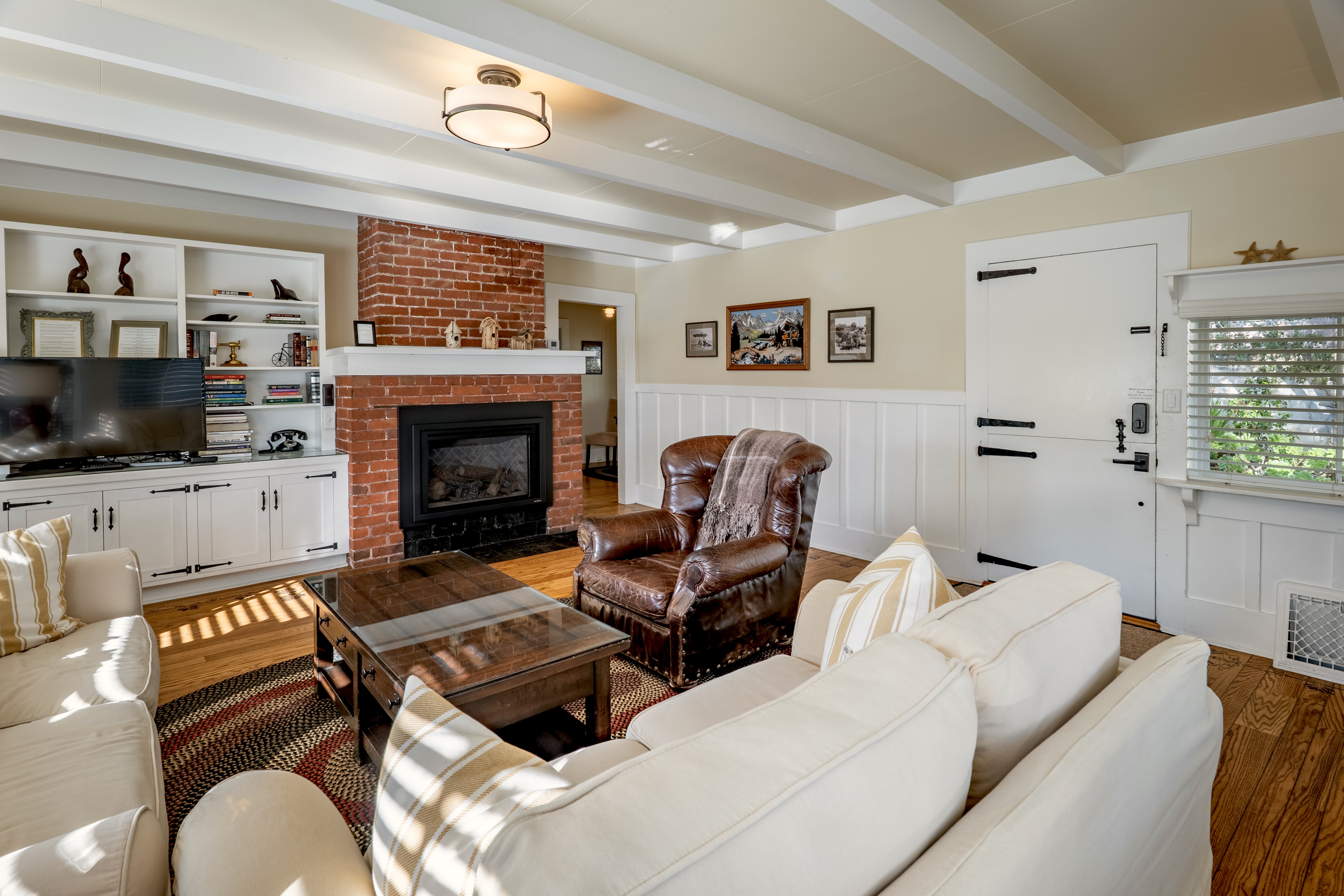 Interior of a cozy living room with white-beamed ceilings, featuring a red brick fireplace with a black insert, white built-in shelving, a leather armchair, and light-colored sofas around a wooden coffee table.