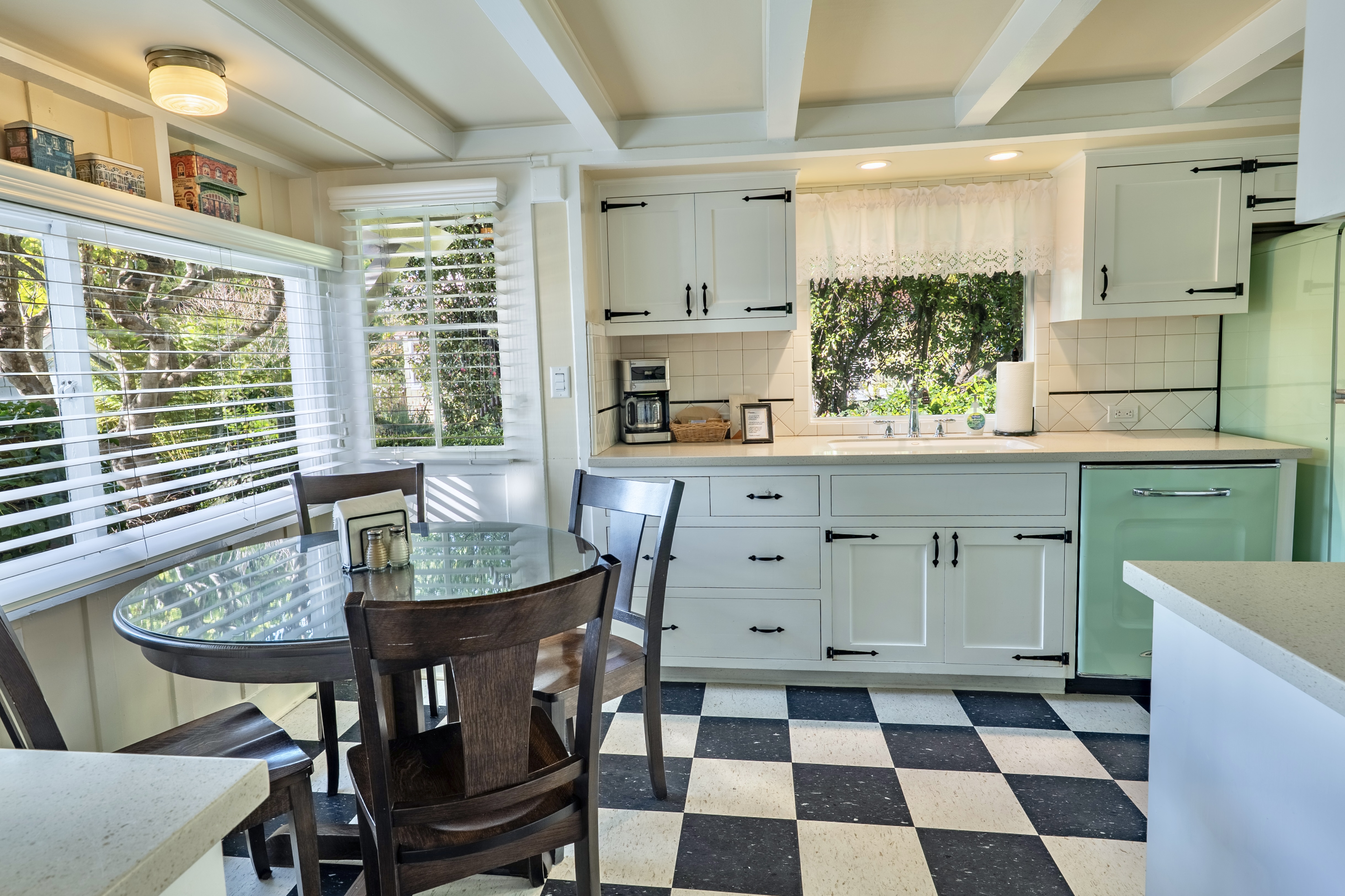 A bright dining area and retro kitchen with black and white checkered floors, featuring a dark wood dining table and chairs next to a large window, and white cabinets with a seafoam green vintage-style refrigerator.