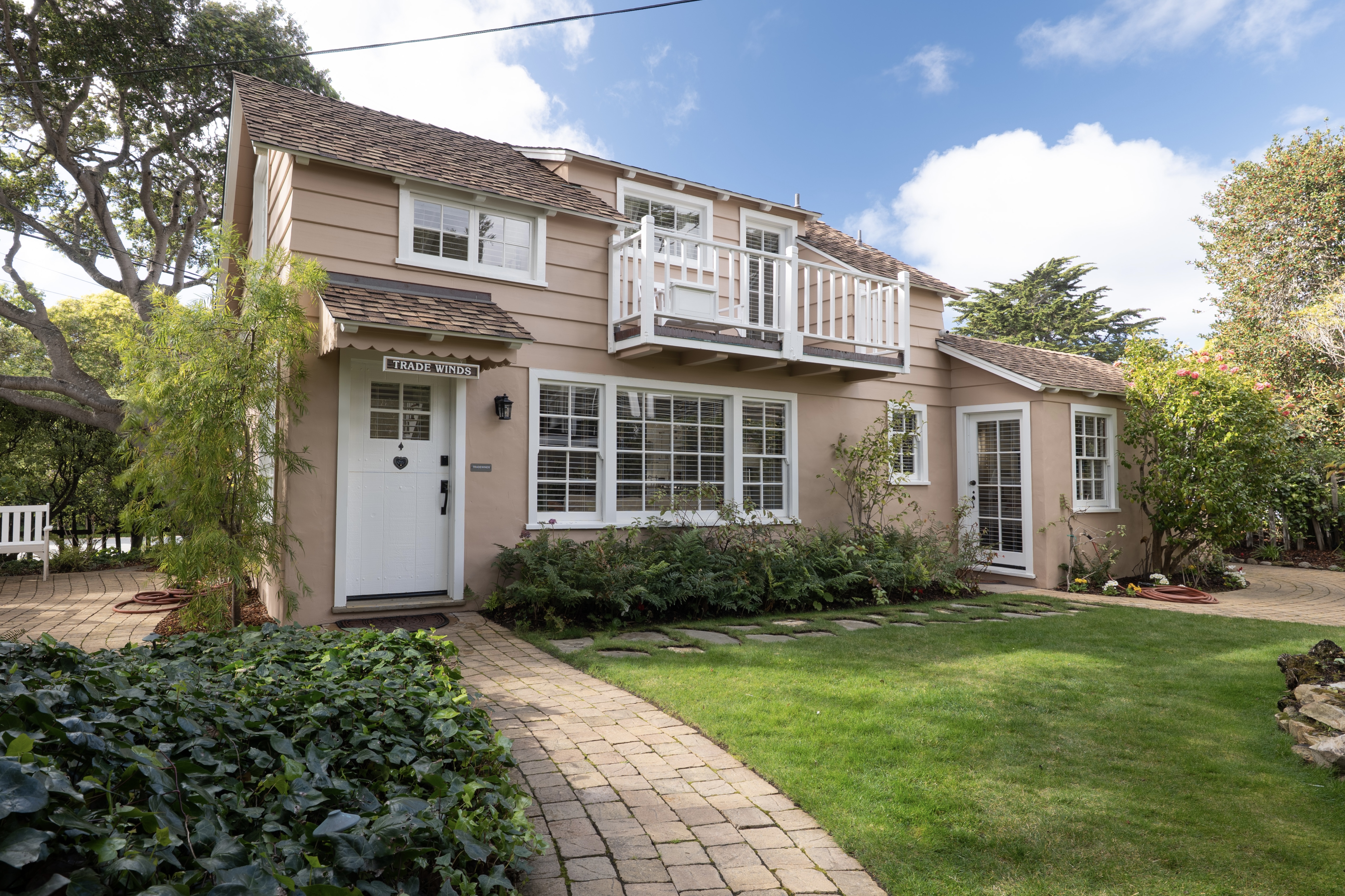 Exterior view of a two-story pinkish tan cottage with a white front door and a second-floor balcony, featuring a paver walkway leading through a green lawn and garden.