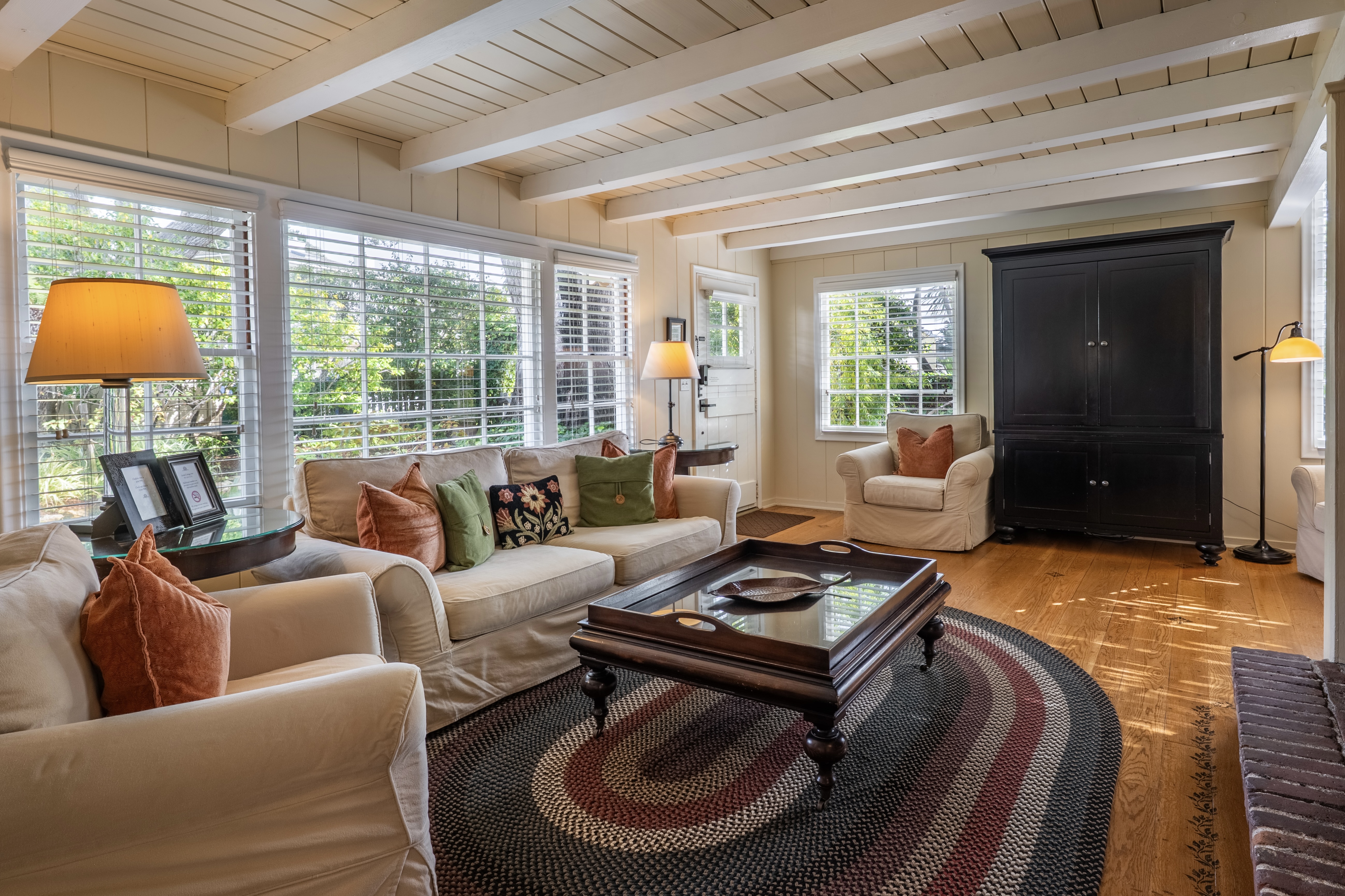 A sunlit living room with white-beamed ceilings and hardwood floors, featuring a large wall of windows, a beige sofa with colorful accent pillows, and a large black armoire.