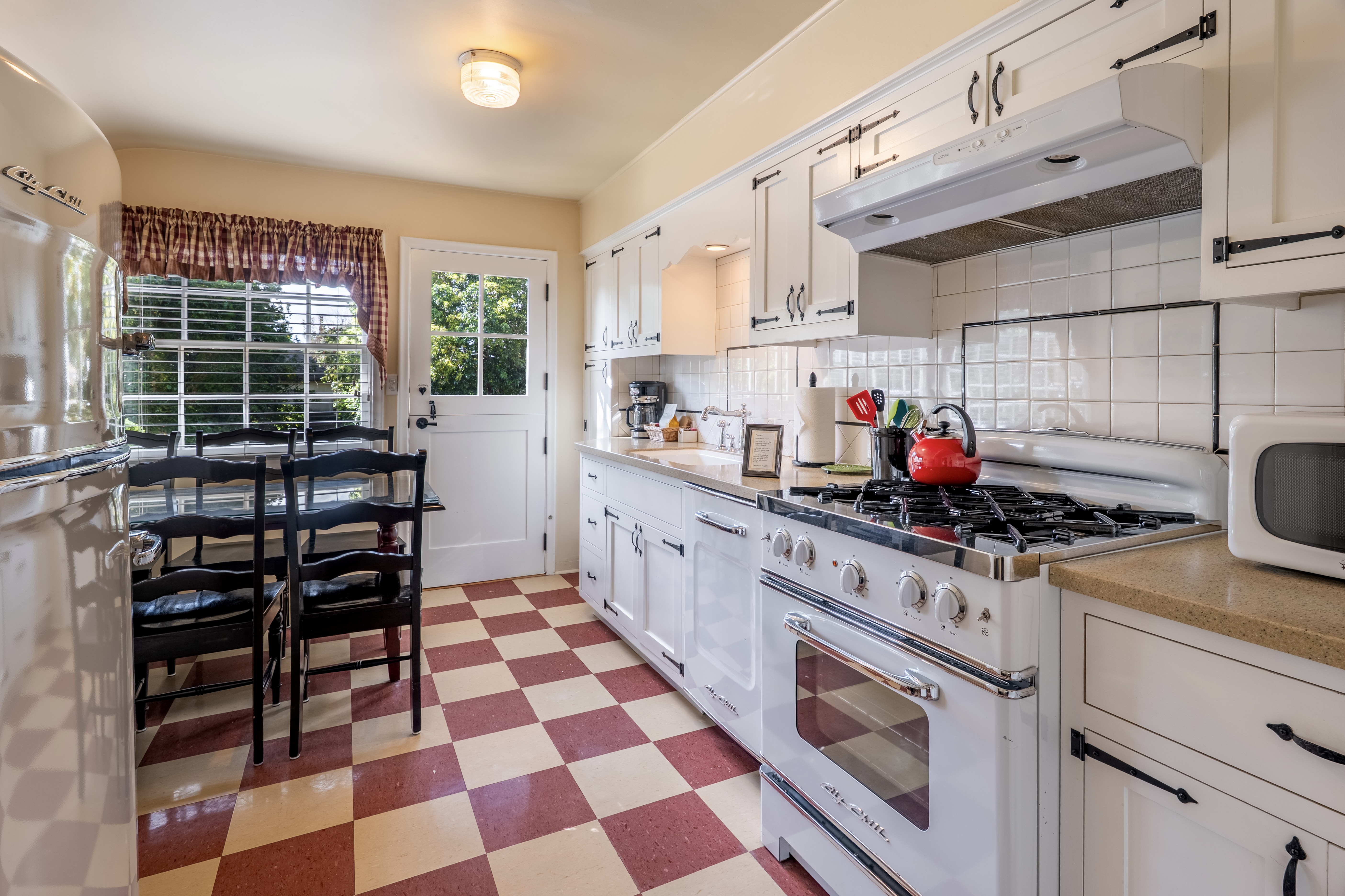 A vintage-style kitchen with a red and white checkered floor, featuring a large white retro gas range, white cabinets with black iron latches, and a  dark dining table with six chairs next to a window.