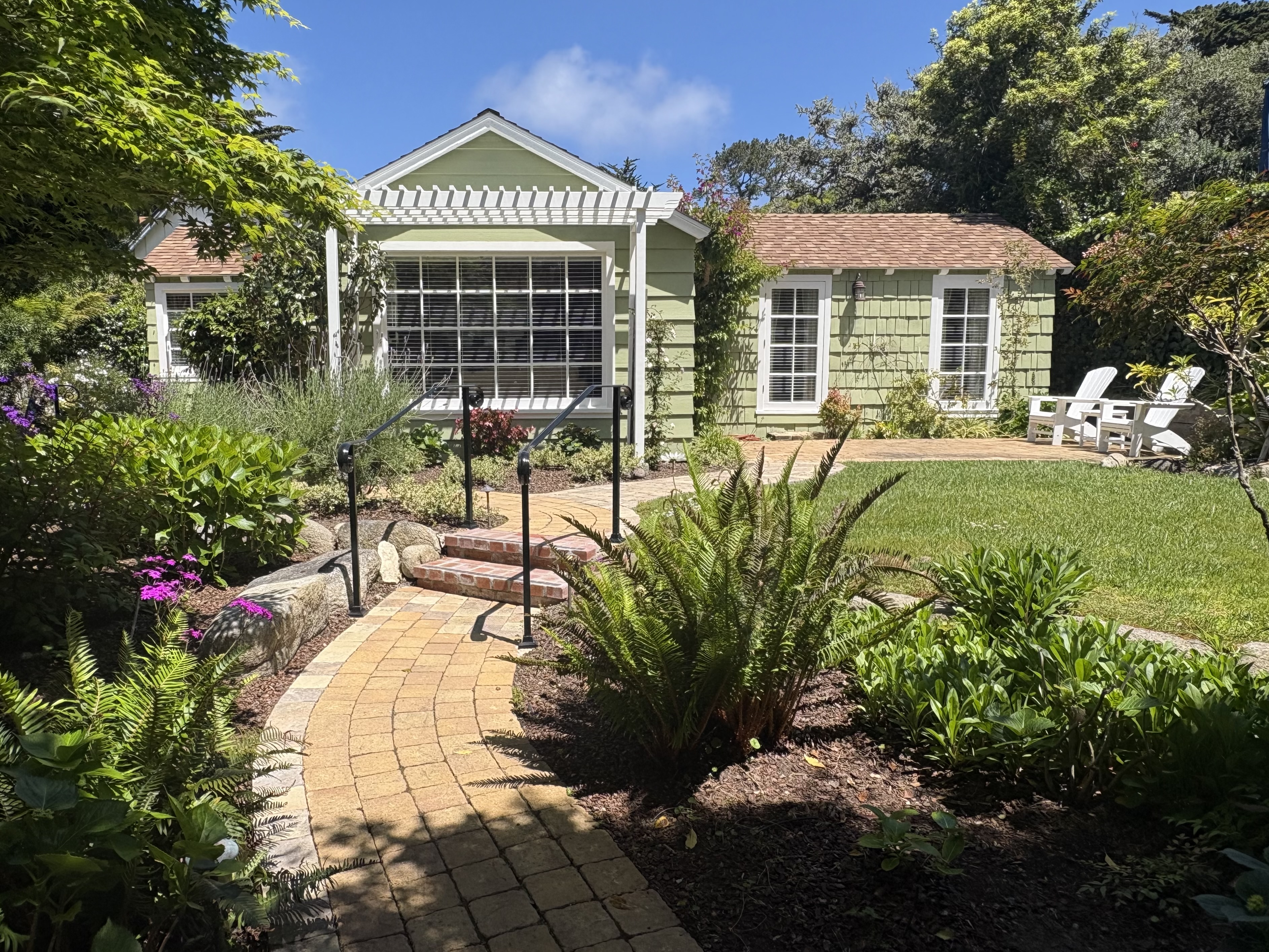Exterior view of a charming pale-green cottage with a gabled roof and white-trimmed windows, featuring a paver path leading up brick steps through a lush green lawn and garden.