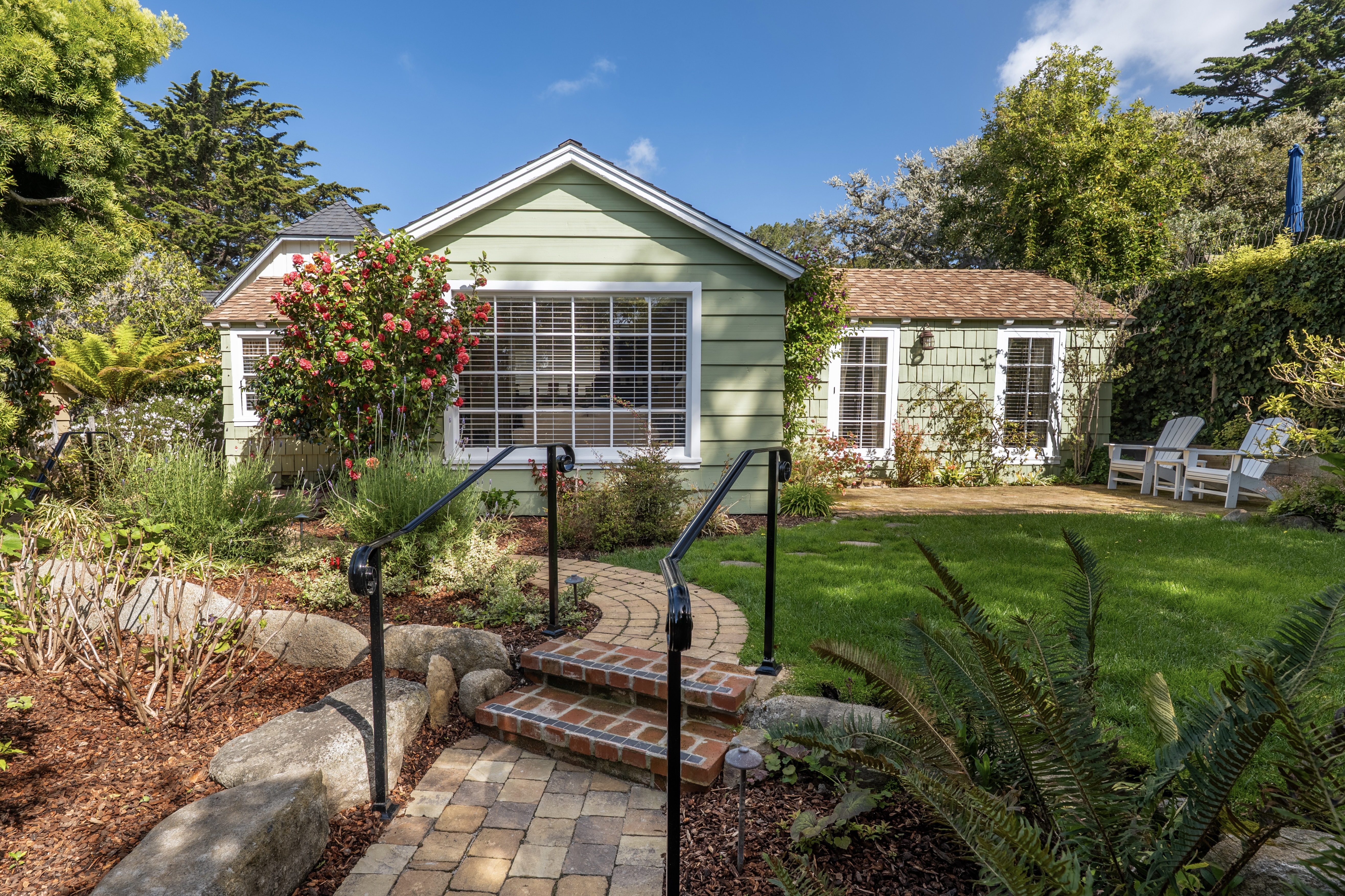 Exterior view of a charming pale-green cottage with a gabled roof and white-trimmed windows, featuring a paver path leading up brick steps through a lush green lawn and garden.