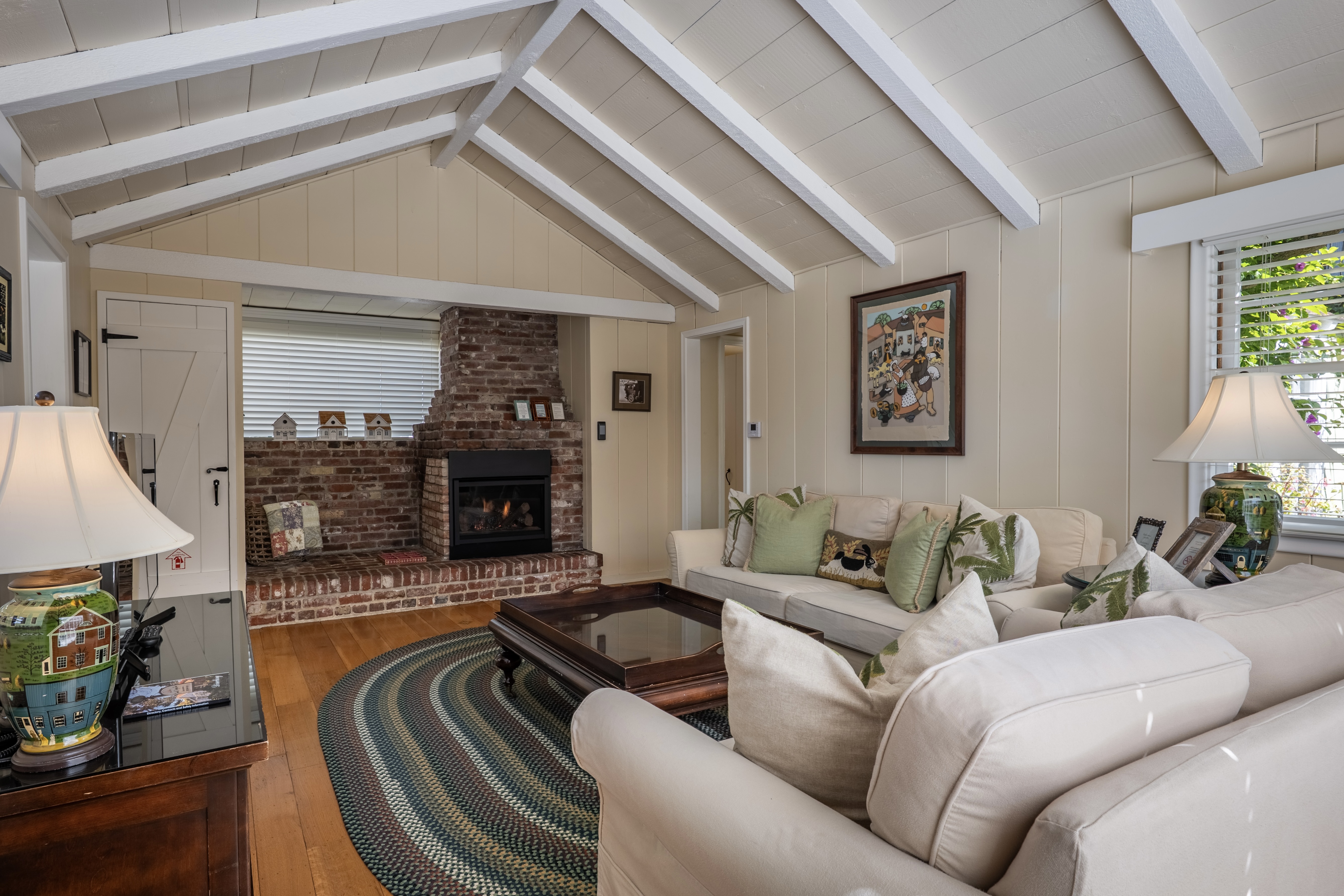 A cozy living room with a vaulted white-beamed ceiling and hardwood floors, featuring a brick,  black insert fireplace, neutral-toned sofas with green accent pillows, and a large oval area rug.