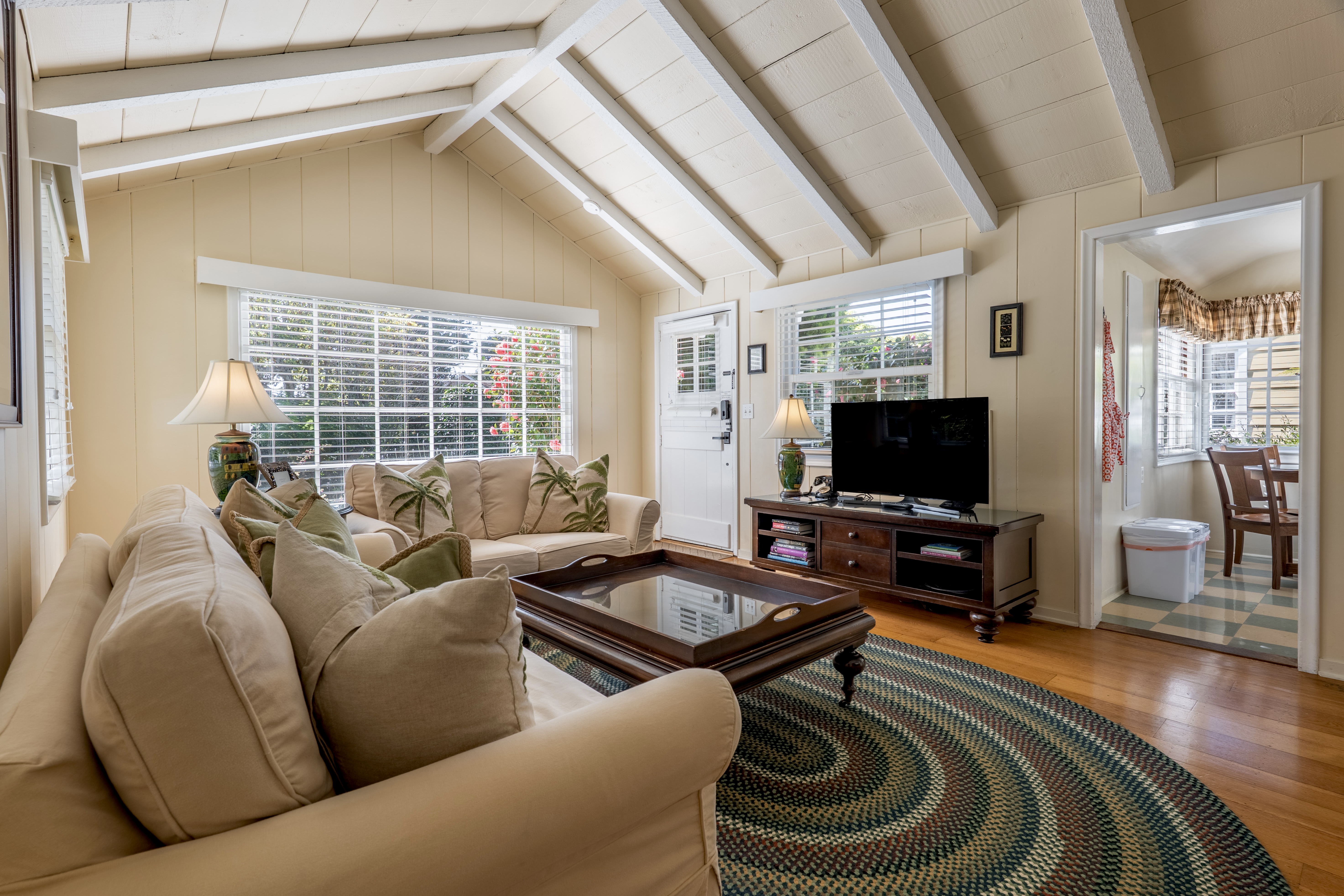 A cozy living room with a vaulted white-beamed ceiling and hardwood floors, a flatscreen TV, neutral-toned sofas with green accent pillows, and a large oval area rug.