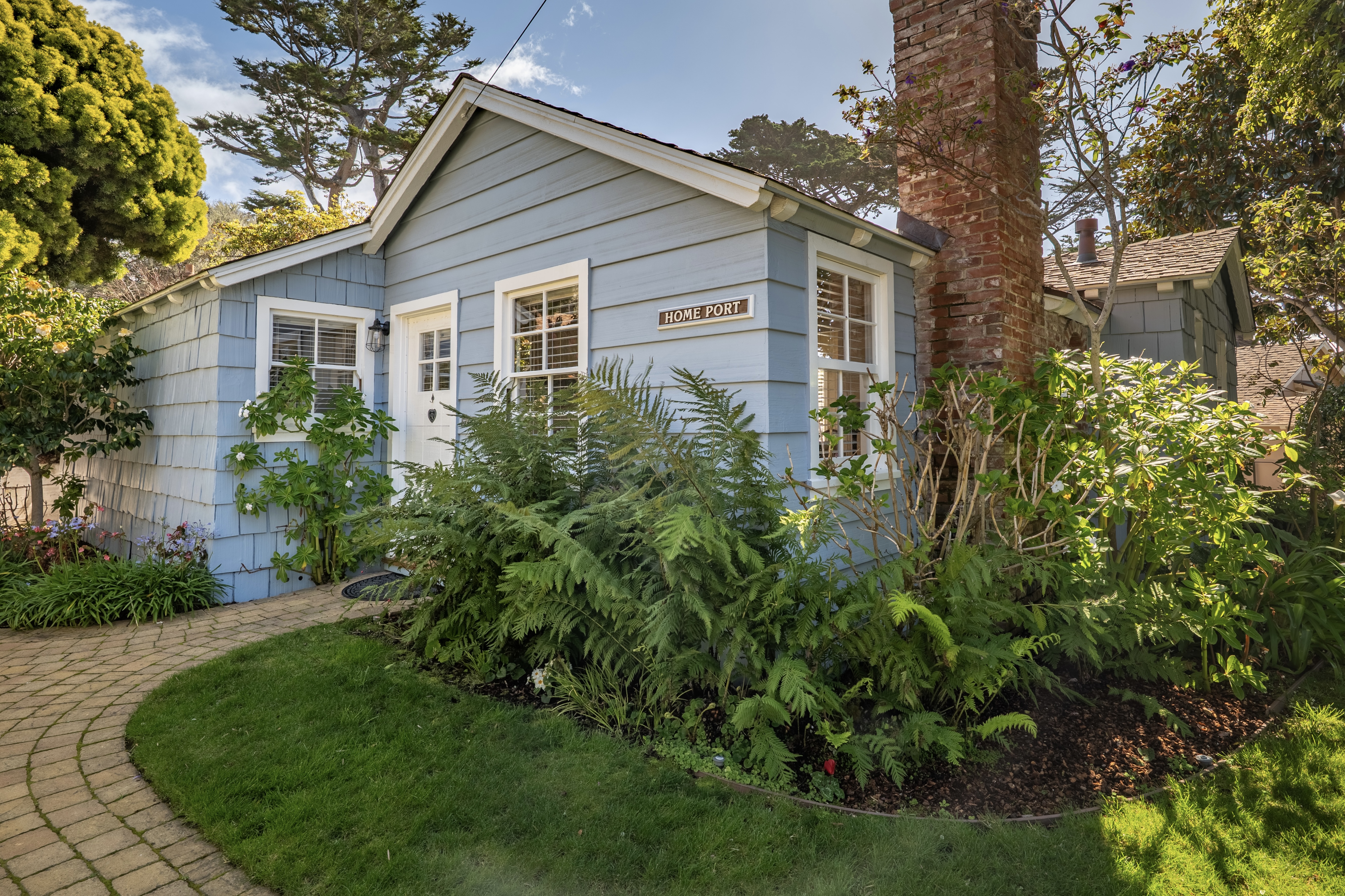 A blue cottage with a white door, surrounded by a lush garden and a winding paver path under a clear blue sky.