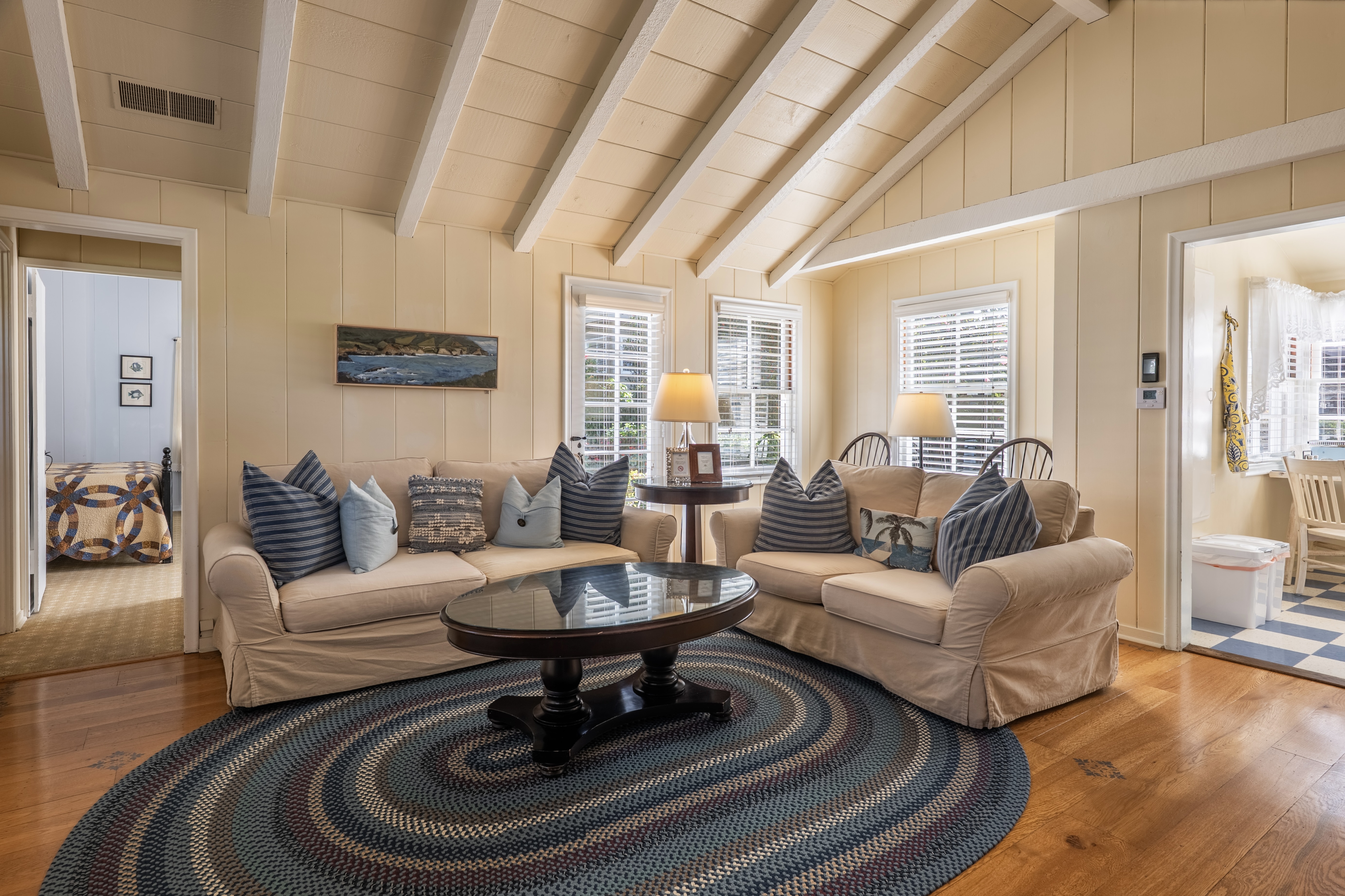 A cozy living room with beige couches, blue throw pillows, white beamed ceiling and a view into the kitchen and the bedroom.