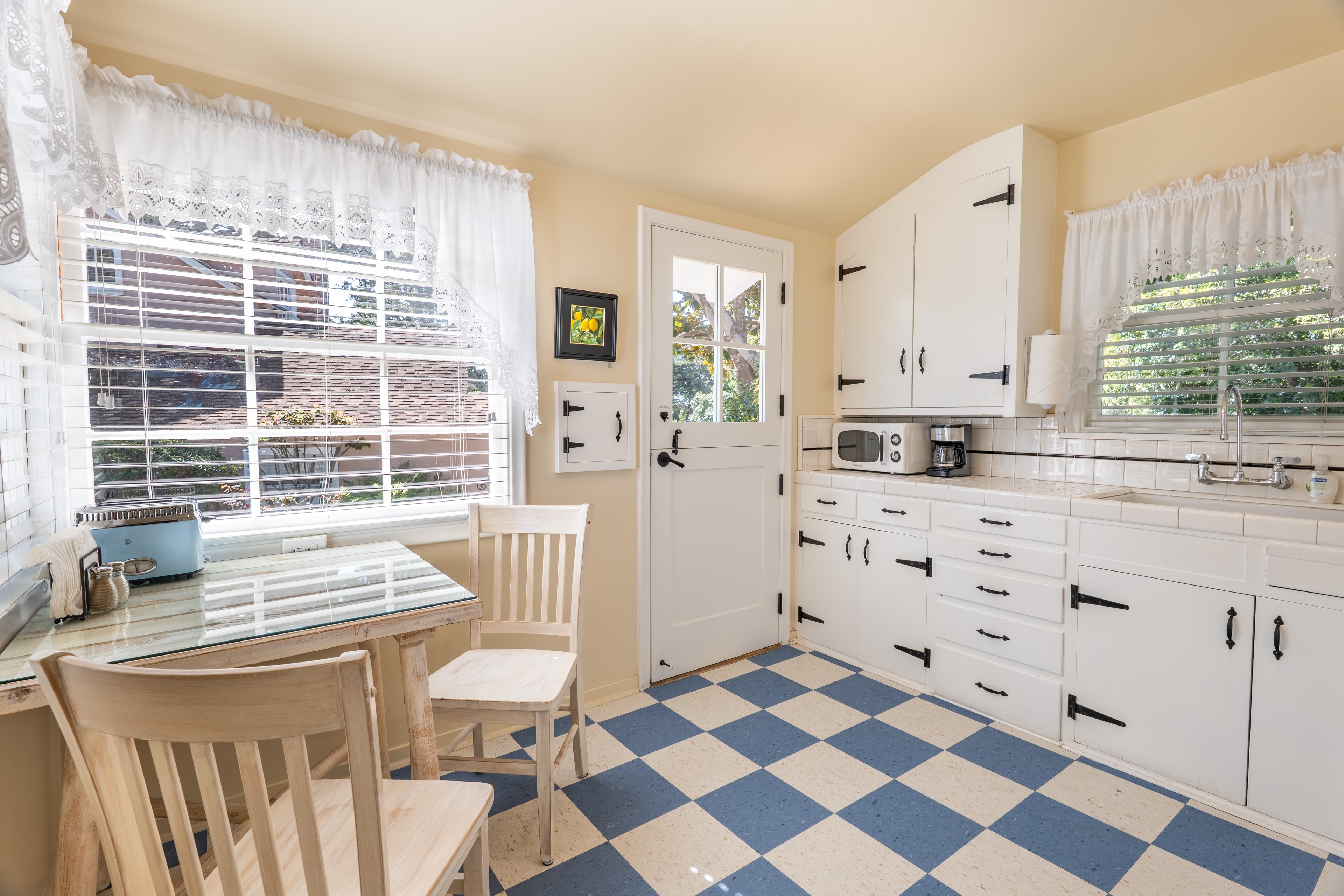 An interior view of a bright, retro-modern kitchen featuring a blue and white checkered floor, white cabinets with black hardware, and a small dining table with two chairs by a window with white lace curtains.