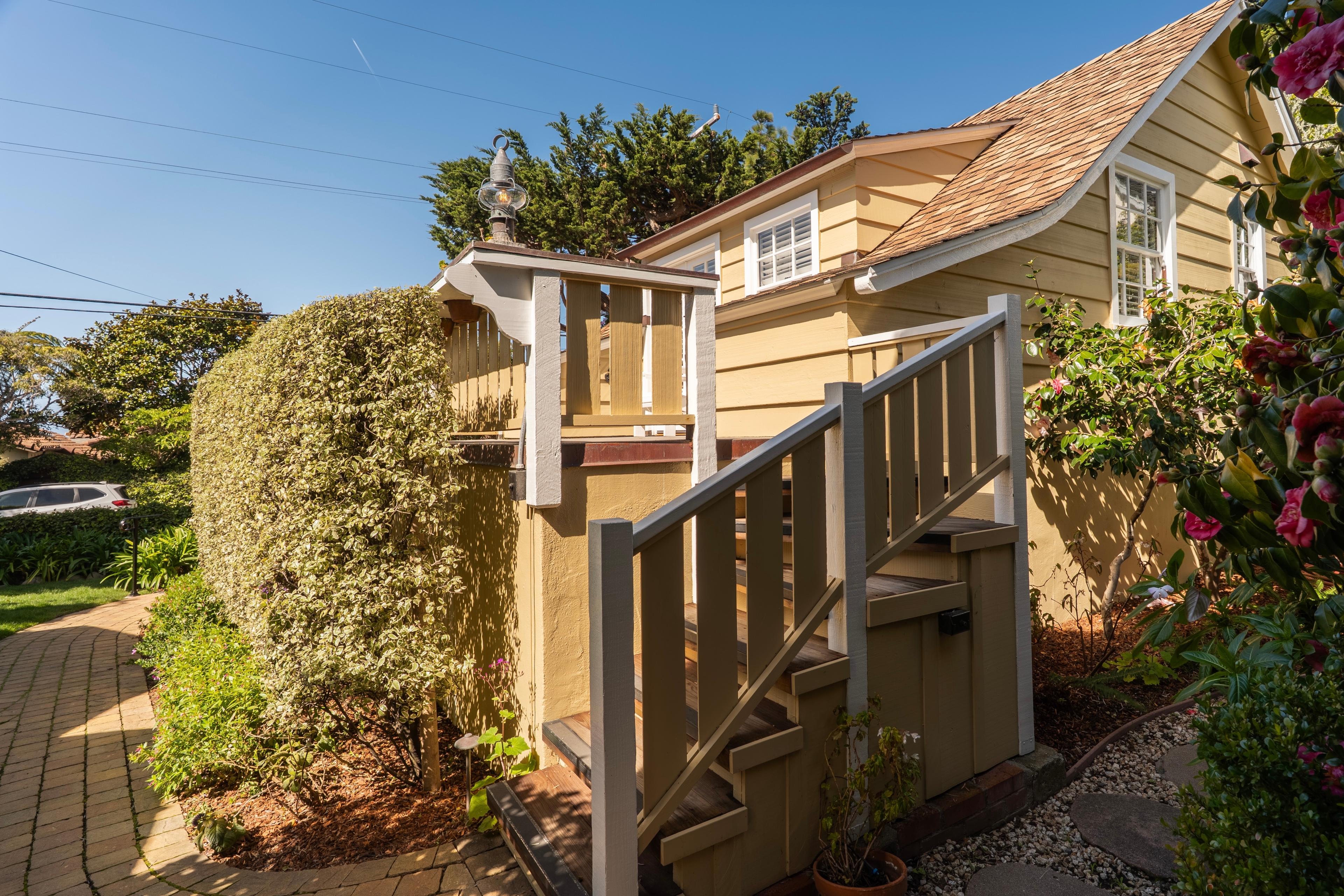 Exterior view of a two-story tan cottage with flower boxes on a second-story wooden balcony, surrounded by lush green bushes.