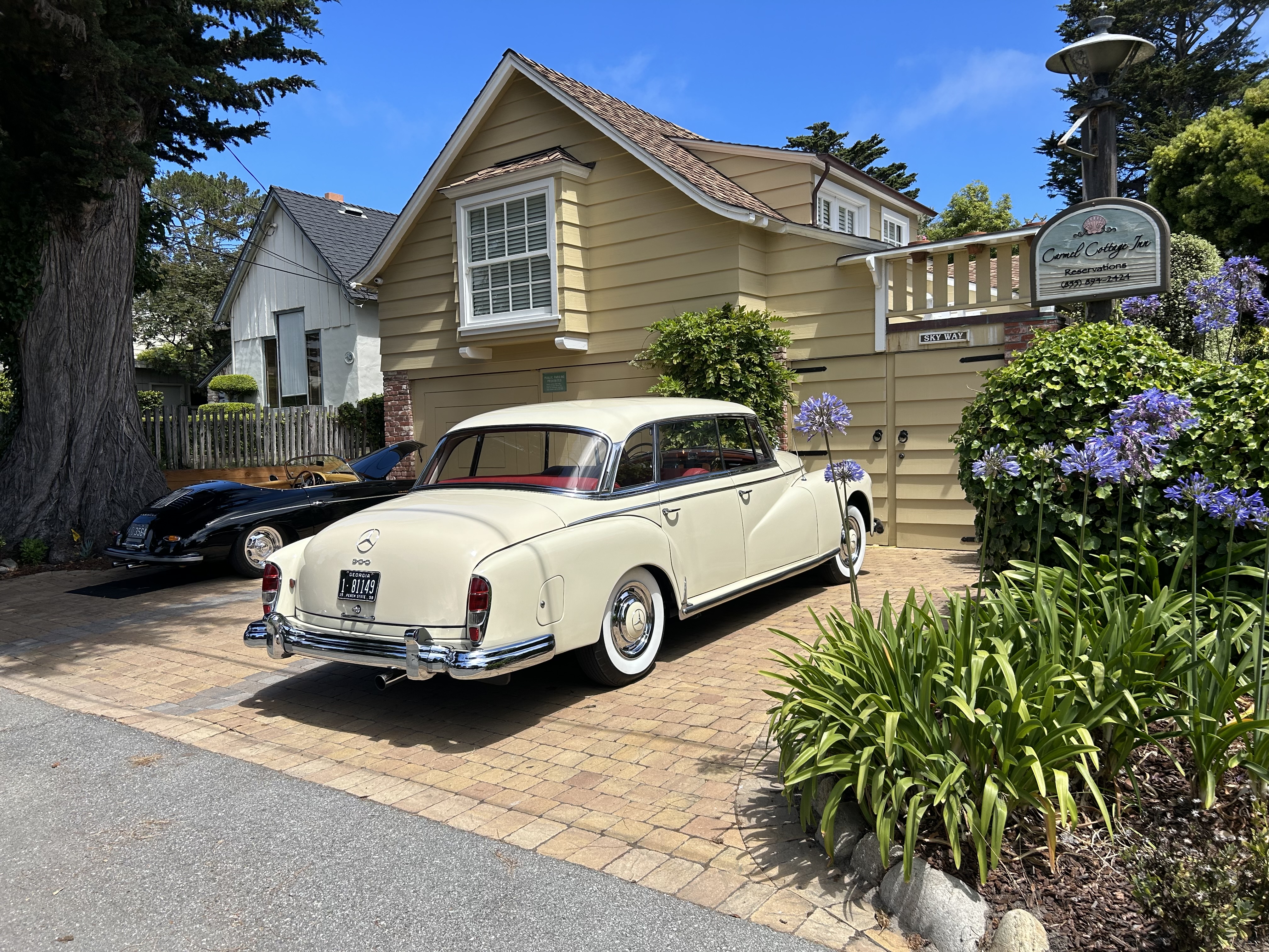 Exterior view of a tan two-story cottage with a wooden balcony and flower boxes, with a vintage cream-colored car parked on the stone driveway in front of the building.