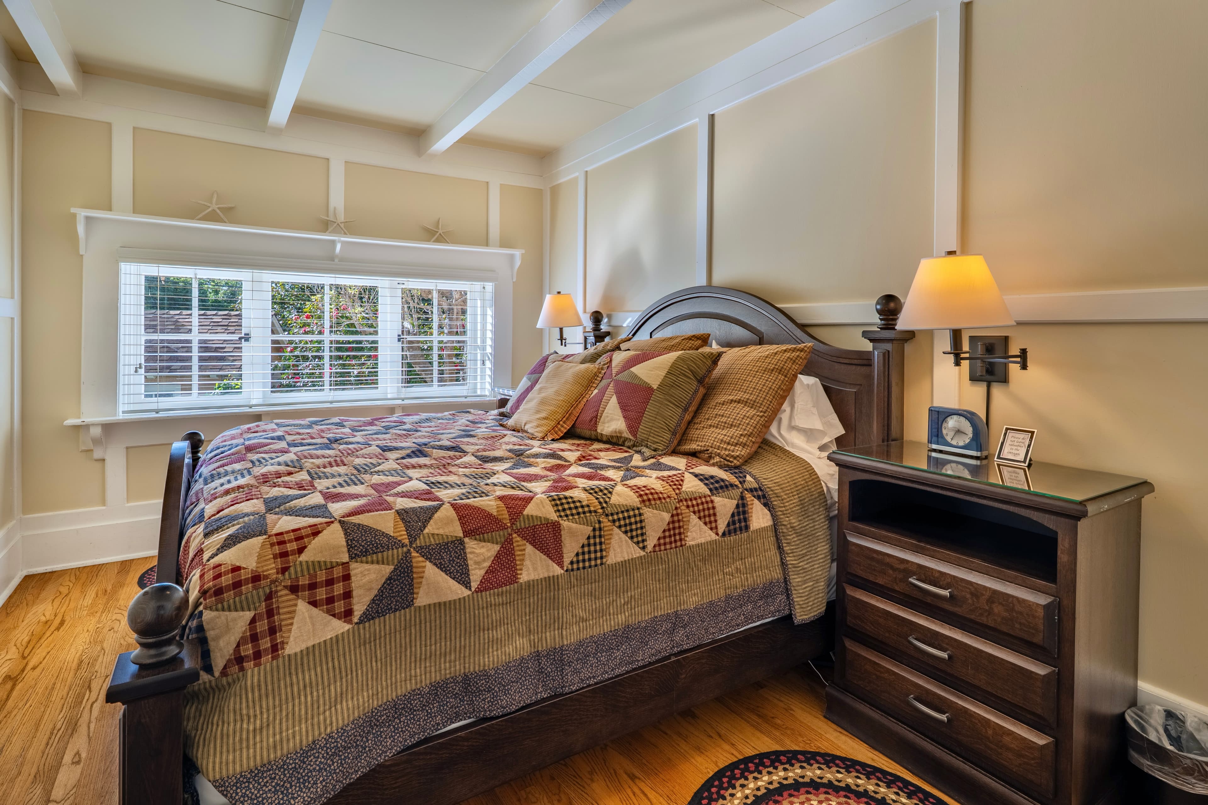 A cozy cottage bedroom featuring a dark wood bed with a patterned quilt, set against beige paneled walls with white trim and a white-beamed ceiling.