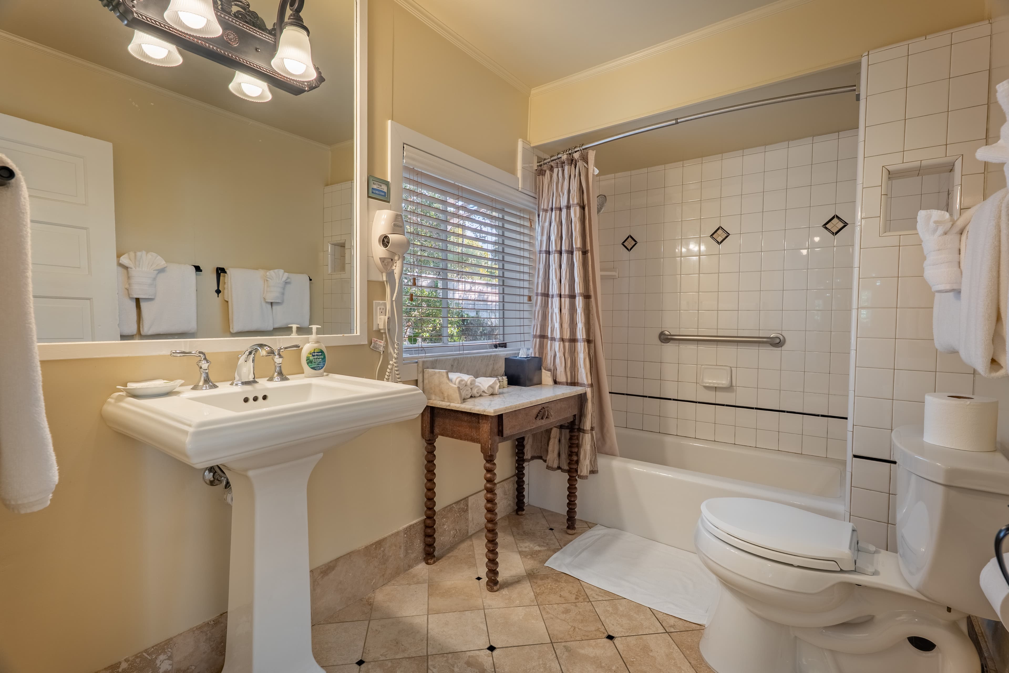 A bathroom with a combination tub/shower, a pedestal sink, a vanity table and a toilet.