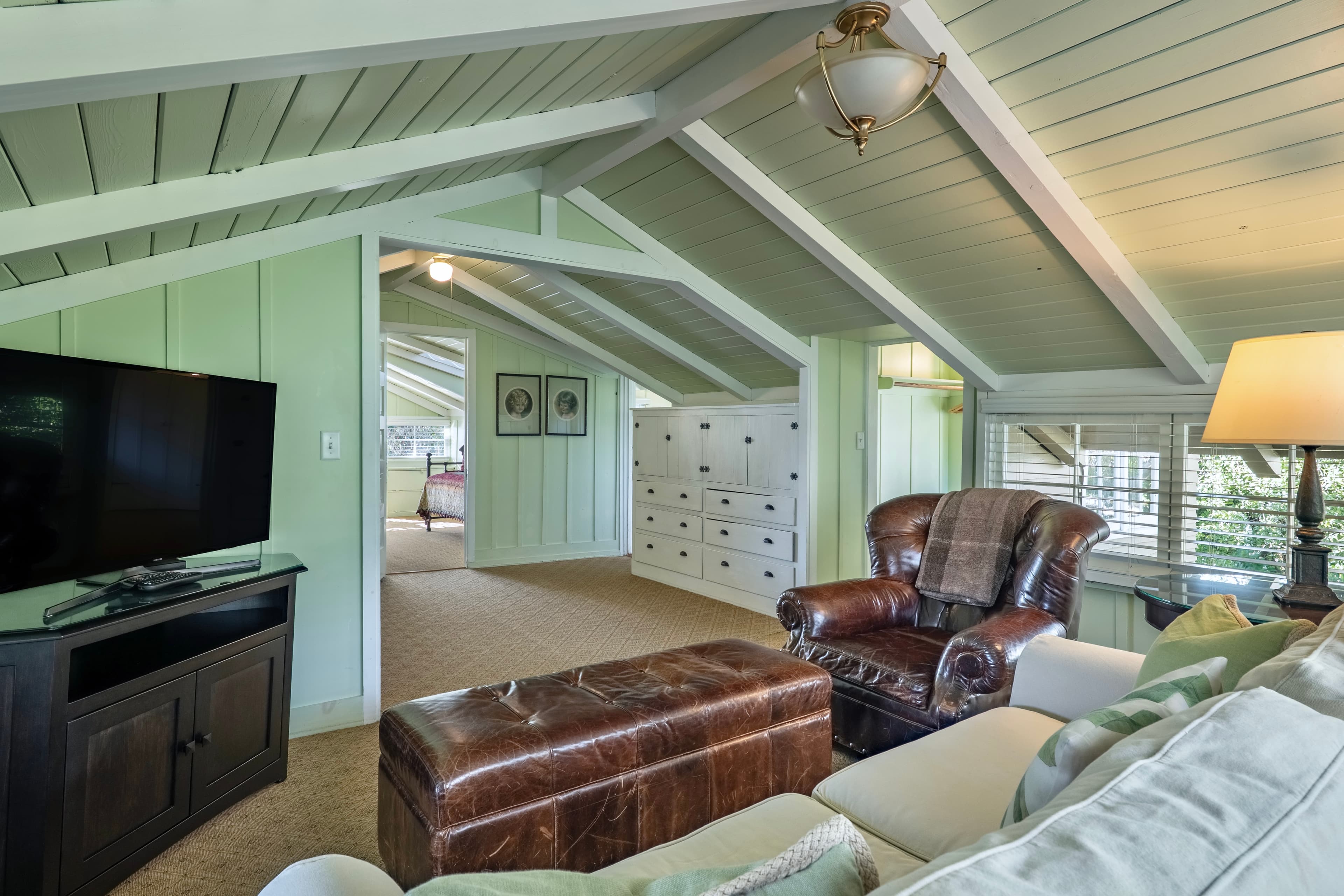 A cozy upstairs living area with a vaulted white-beamed ceiling, featuring a flat-screen TV on a dark wood stand, a leather armchair with a tufted ottoman, and a glimpse into an adjacent bedroom.