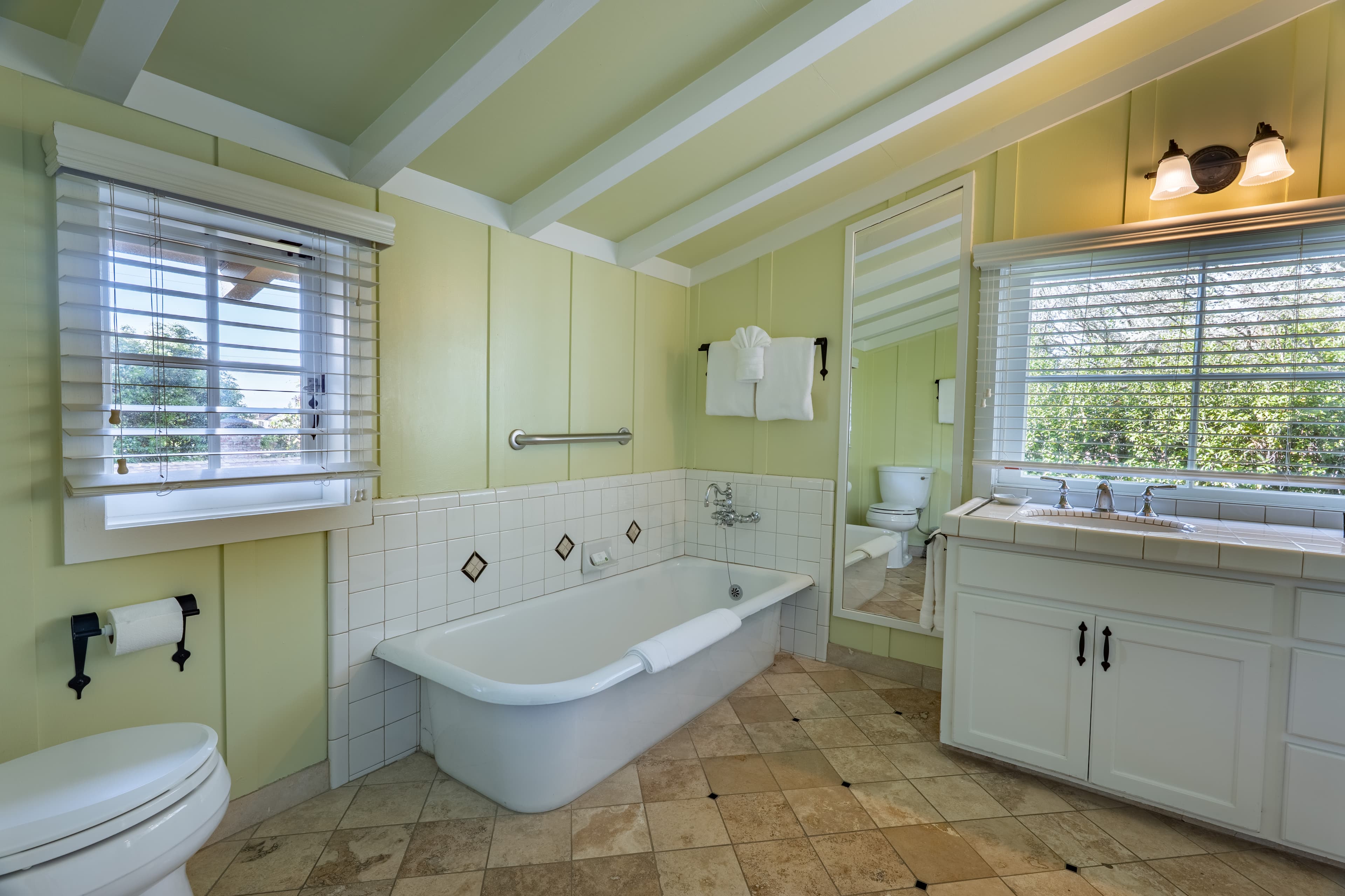 A bright, spacious bathroom with light green walls and a white-beamed ceiling, featuring a large white soaking tub, a wide vanity with double sinks, and a large window with white blinds.