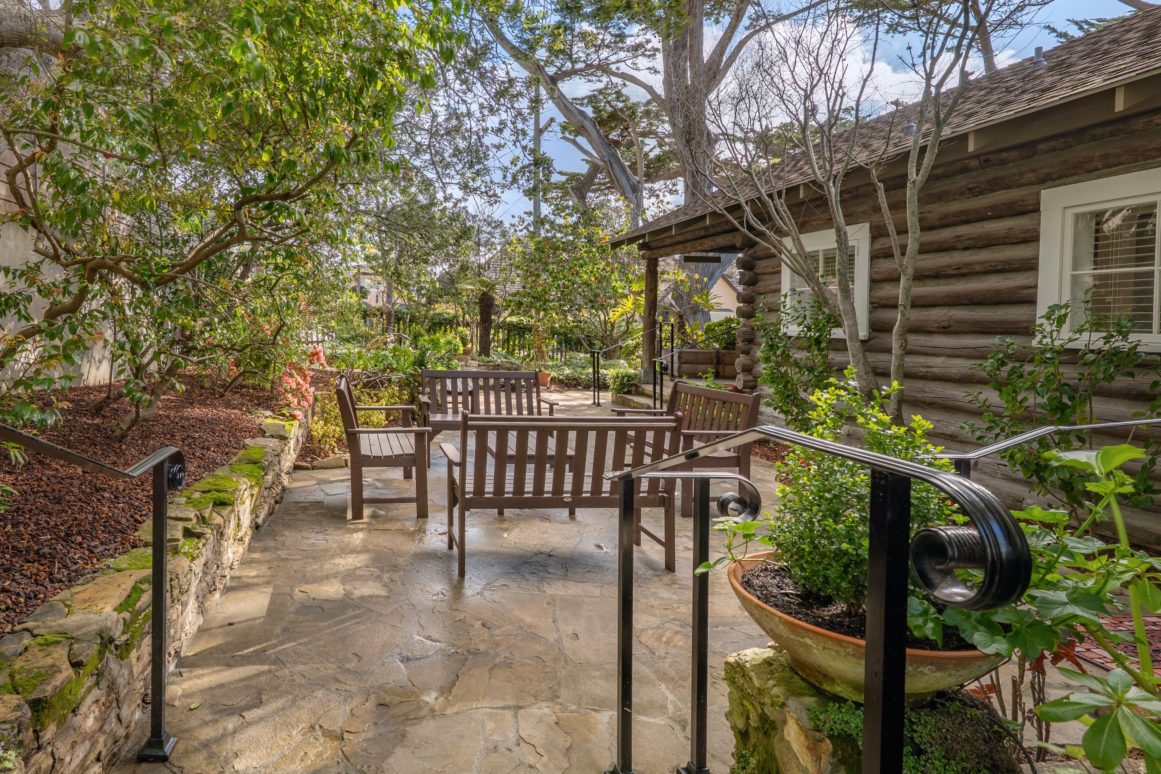 A wide stone patio next to the Log Haven cabin, featuring dark polywood benches and chairs surrounded by lush trees and greenery under a clear blue sky.