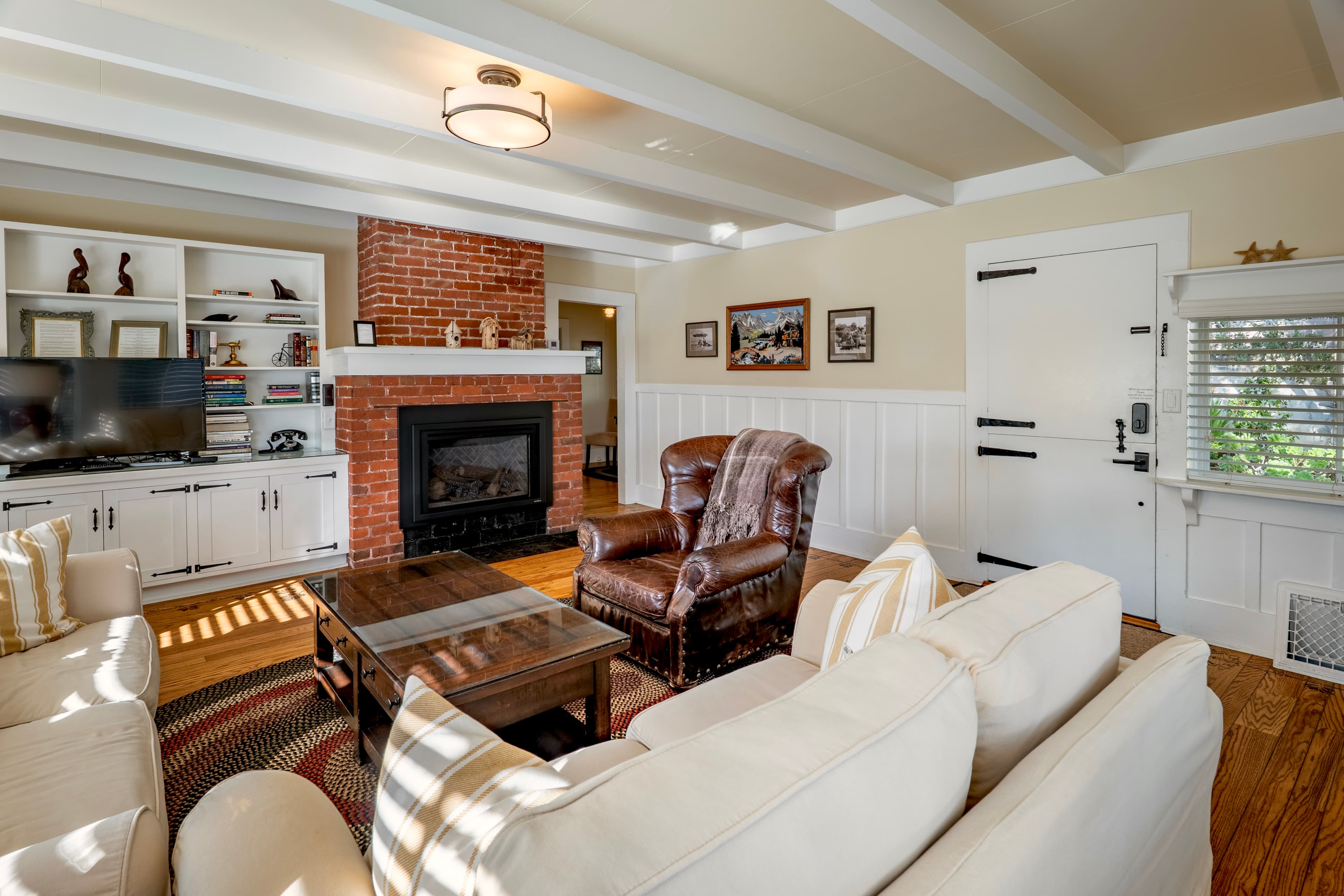 Interior of a cozy living room with white-beamed ceilings, featuring a red brick fireplace with a black insert, white built-in shelving, a leather armchair, and light-colored sofas around a wooden coffee table.