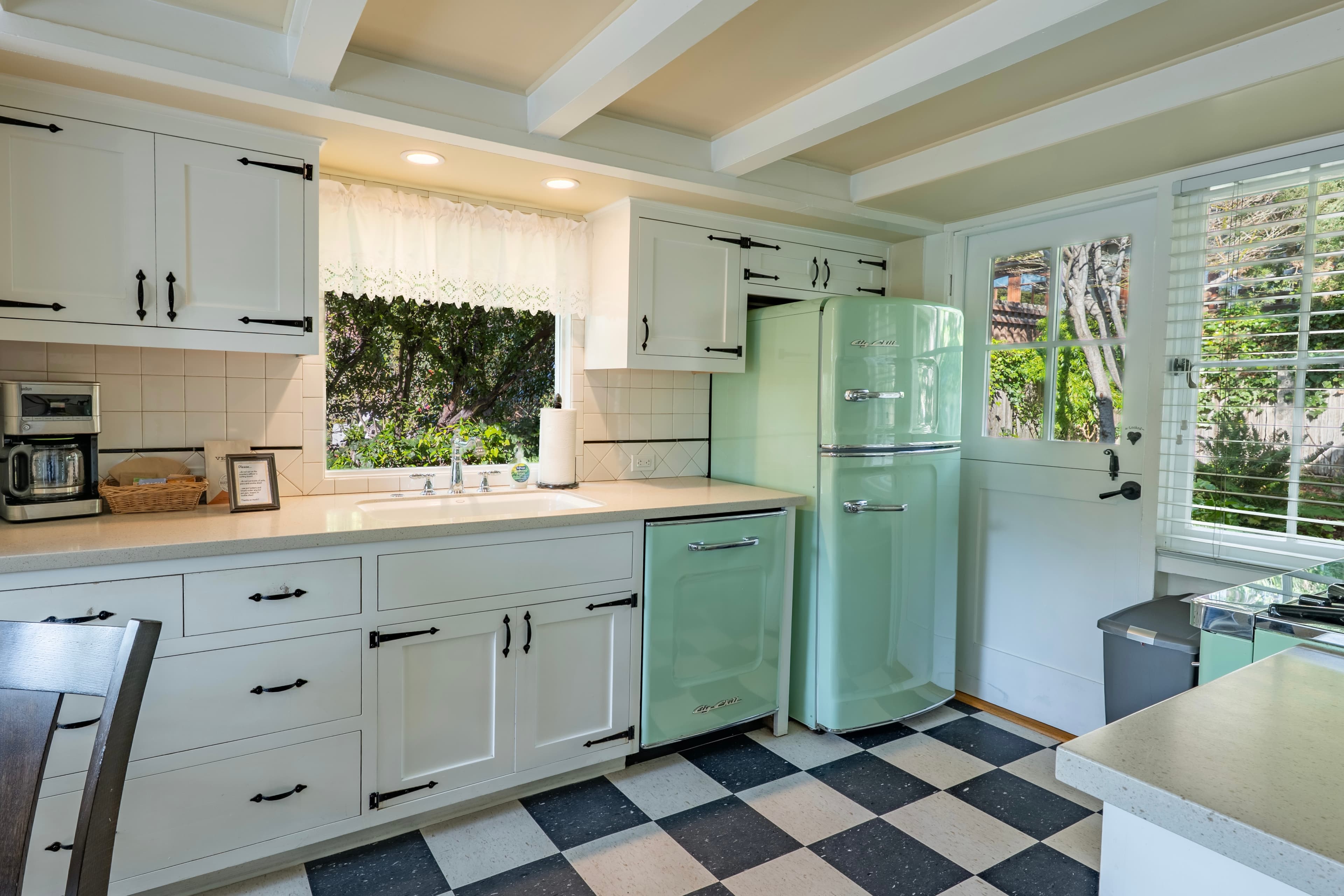 A bright, retro-inspired kitchen with a black and white checkered floor, featuring a seafoam green retro refrigerator and matching dishwasher alongside white cabinetry with black hardware.