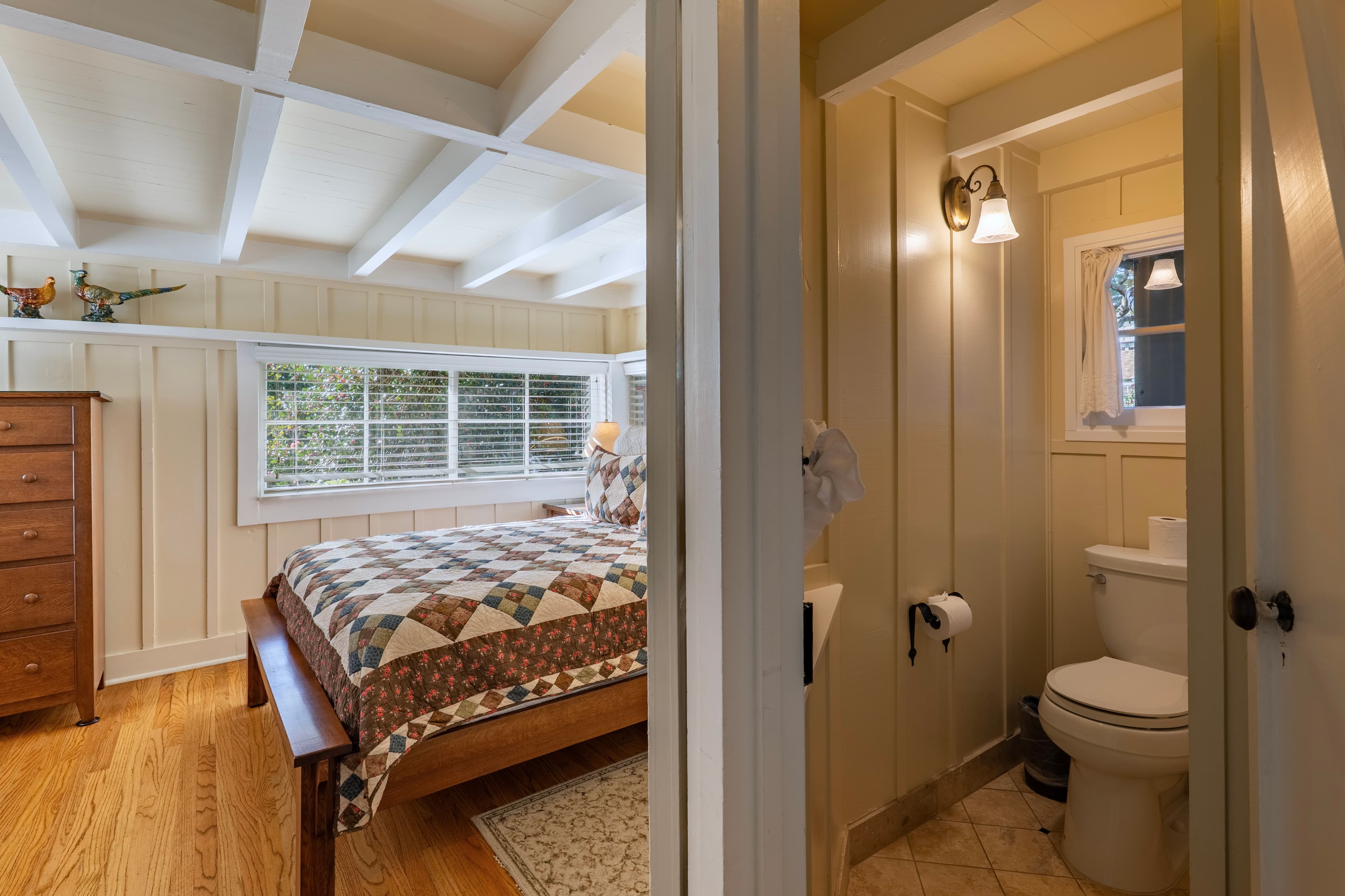 A cozy cottage bedroom with wood floors and beige paneled walls, featuring a wooden bed with a patterned quilt and a tall dresser, with a partial view of an adjacent bathroom.
