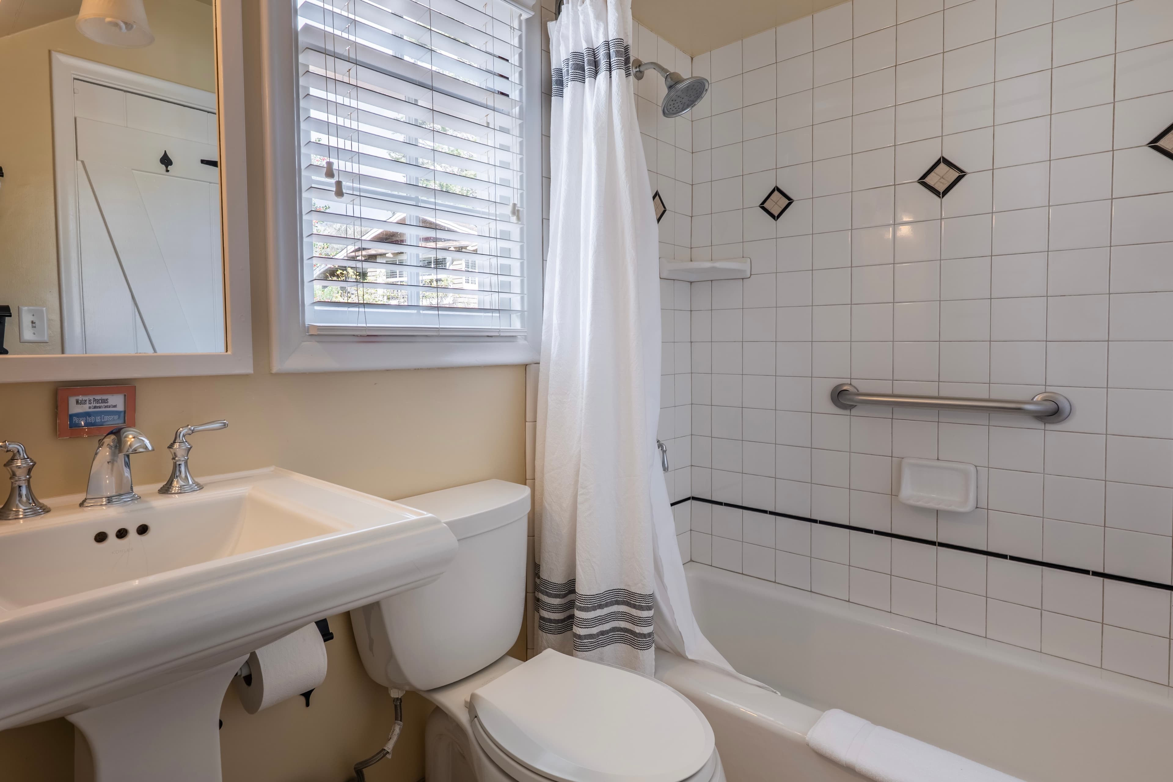 A classic bathroom featuring a white pedestal sink and a white toilet next to a bathtub and shower with white square tiling and decorative black diamond accents.