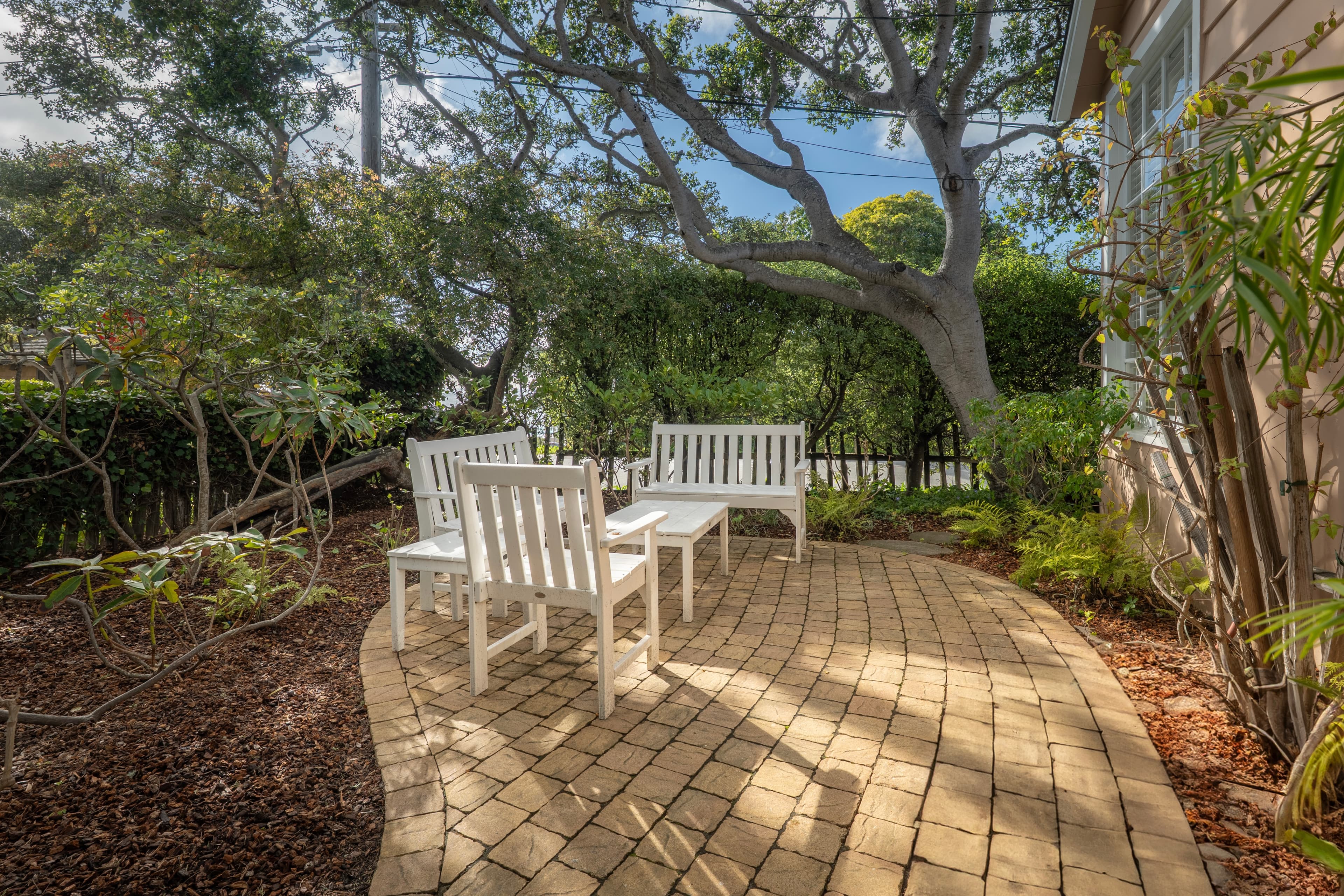 A sunny paver patio with 2 white benches, a matching chair and 2 tables arranged in a cozy seating area, shaded by a large tree with winding branches.
