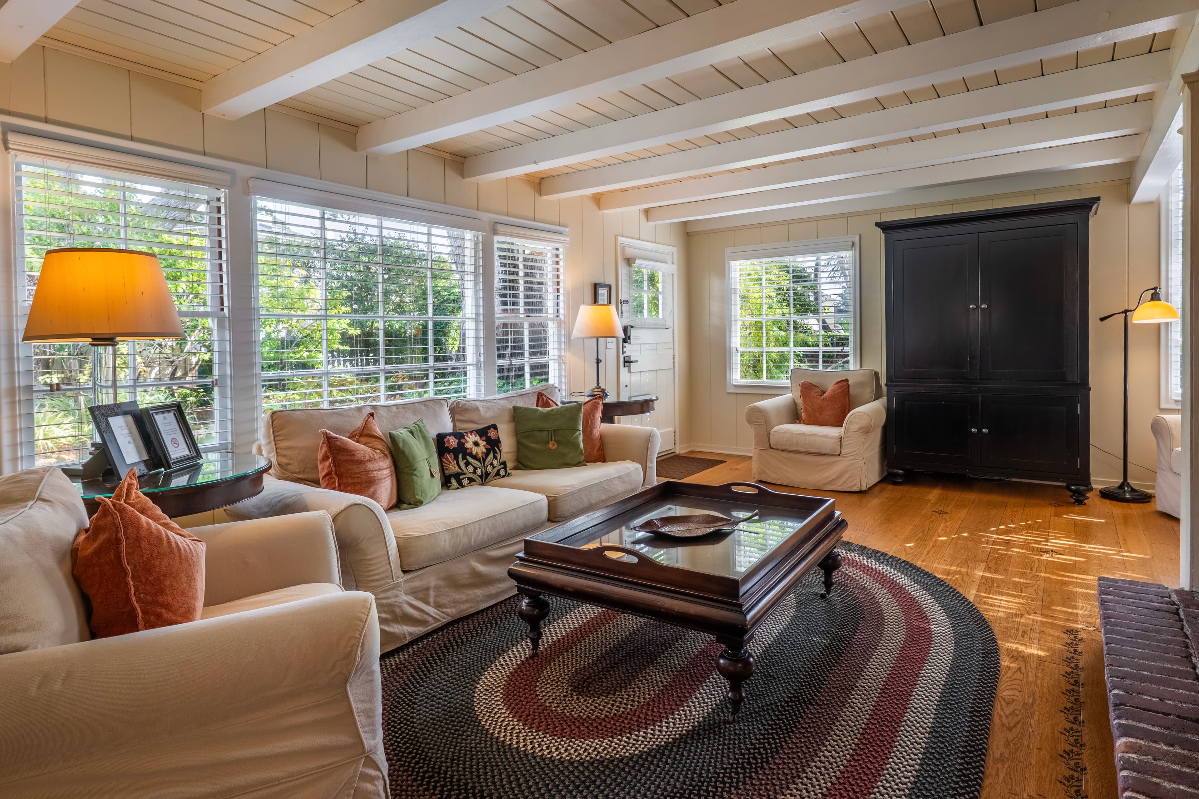 A sunlit living room with white-beamed ceilings and hardwood floors, featuring a large wall of windows, a beige sofa with colorful accent pillows, and a large black armoire.