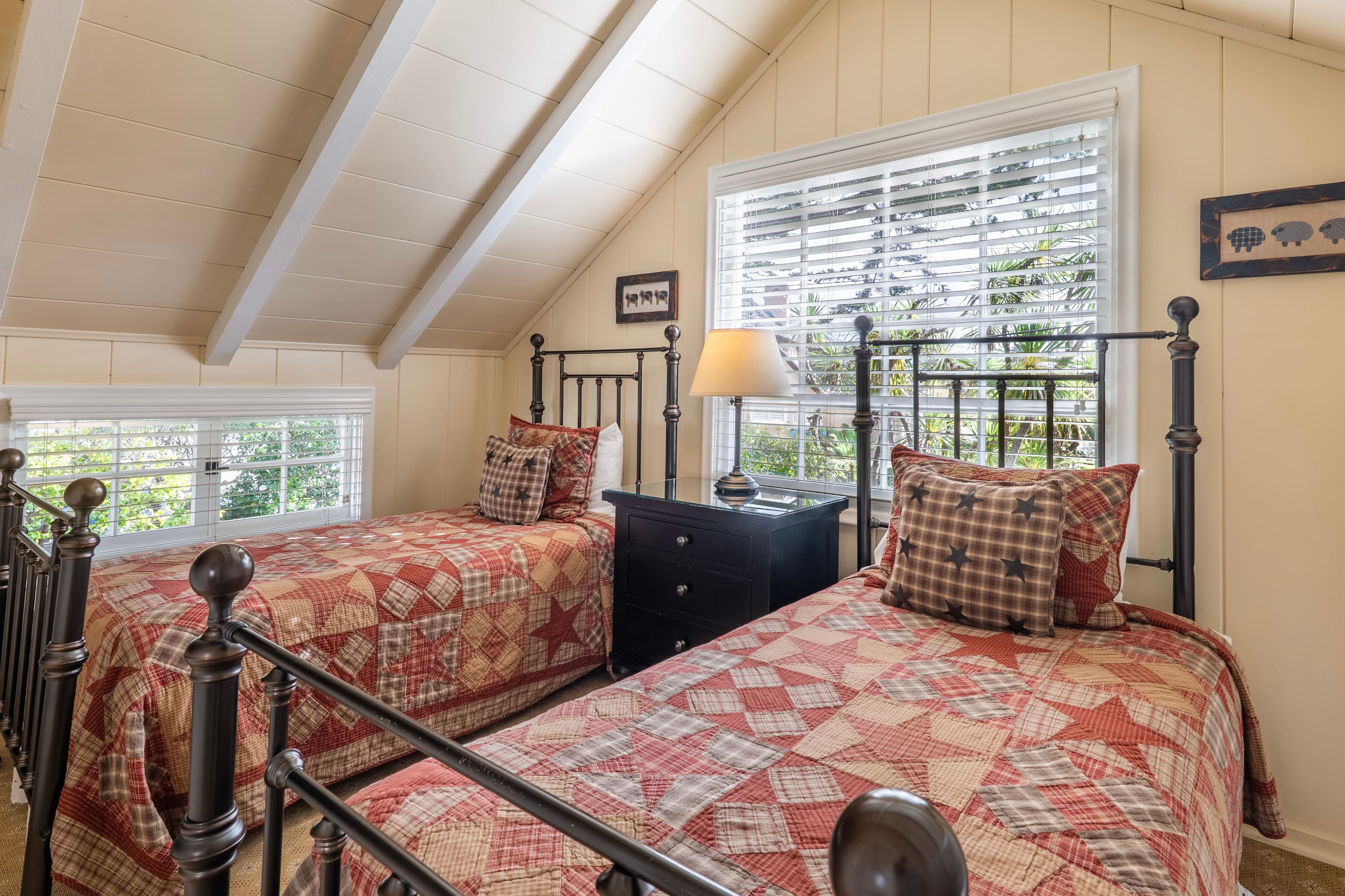 A loft featuring two twin-sized metal bed frames with patterned quilts, set against cream-colored walls under a vaulted white-beamed ceiling.