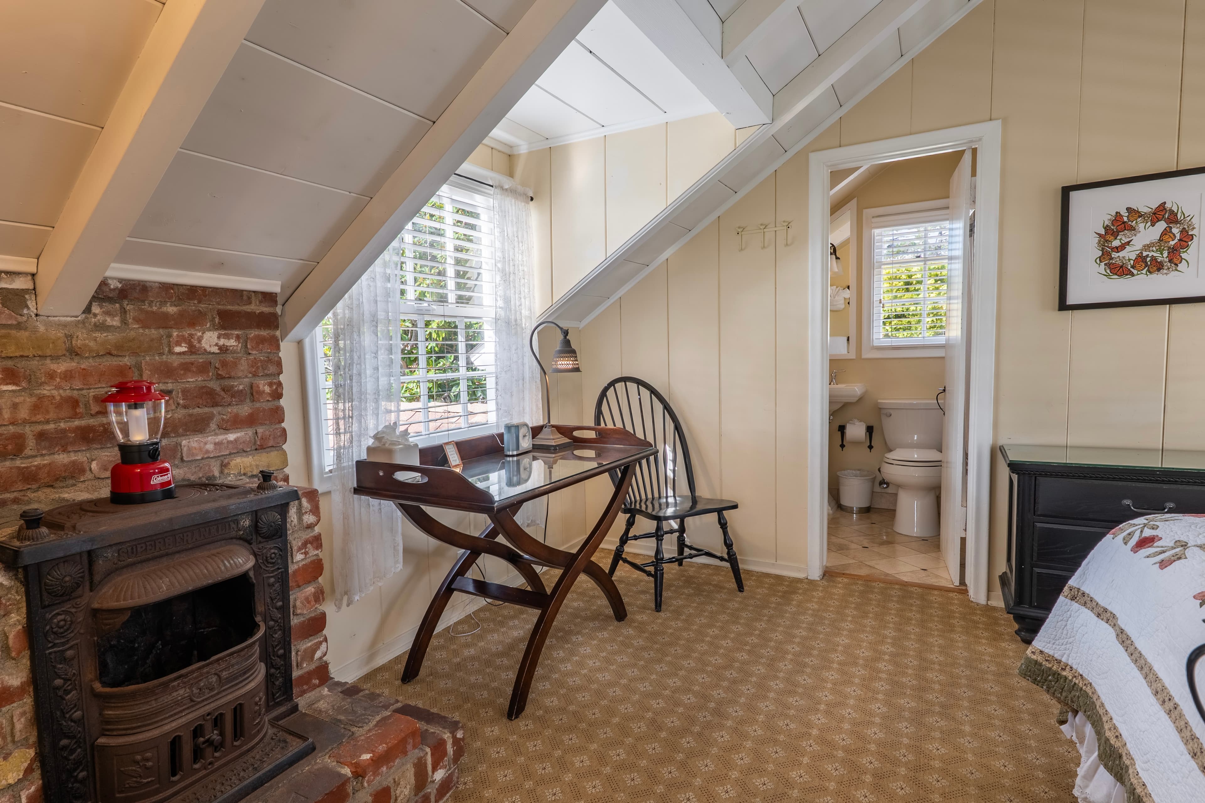 A cozy cottage bedroom featuring a sloped white-beamed ceiling, a decorative brick fireplace, and a dark wood tray-top desk with a chair.
