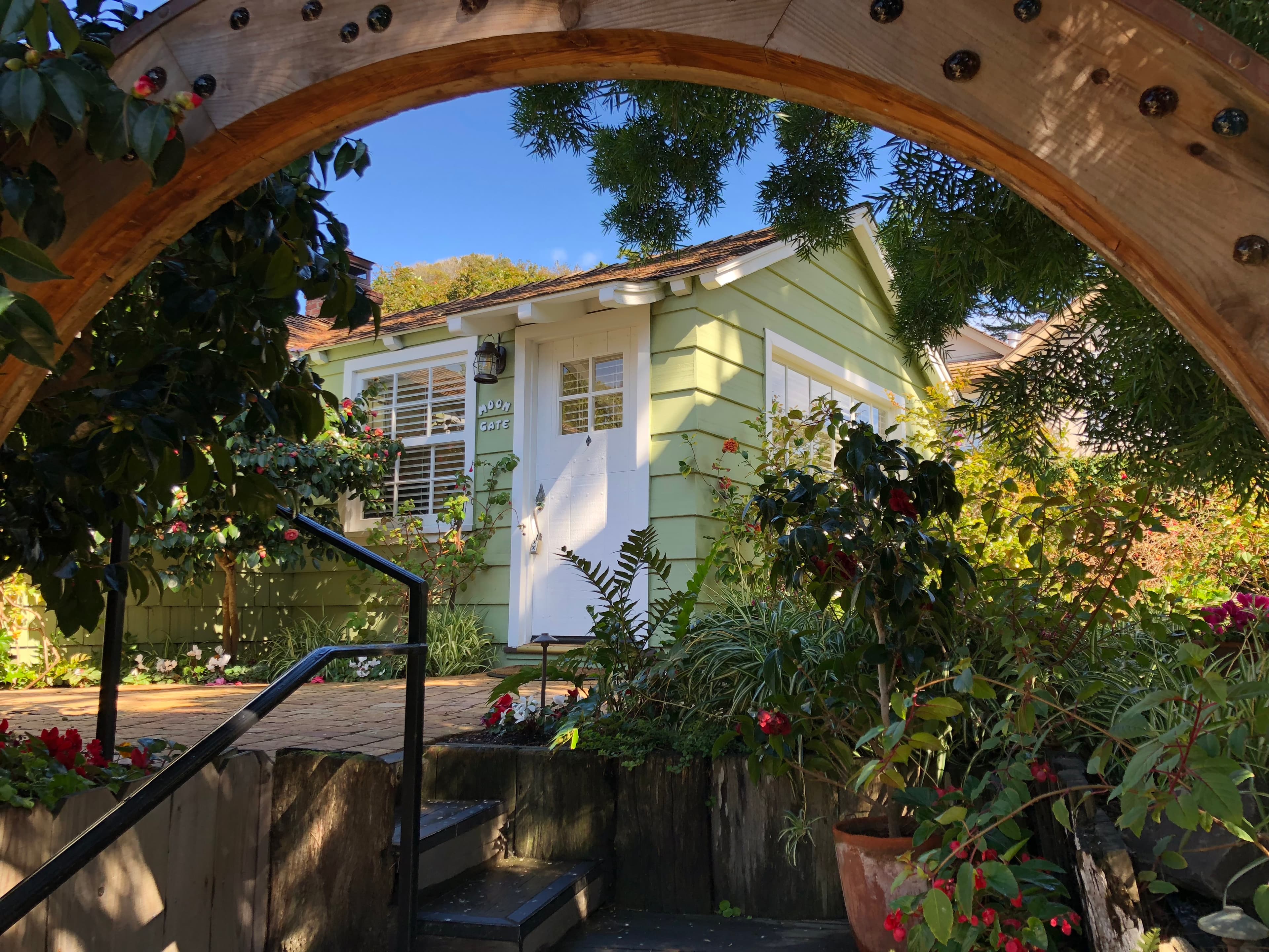 Exterior view of a small, light-green cottage with a white door and windows, partially framed by a rustic wooden archway and surrounded by lush greenery.