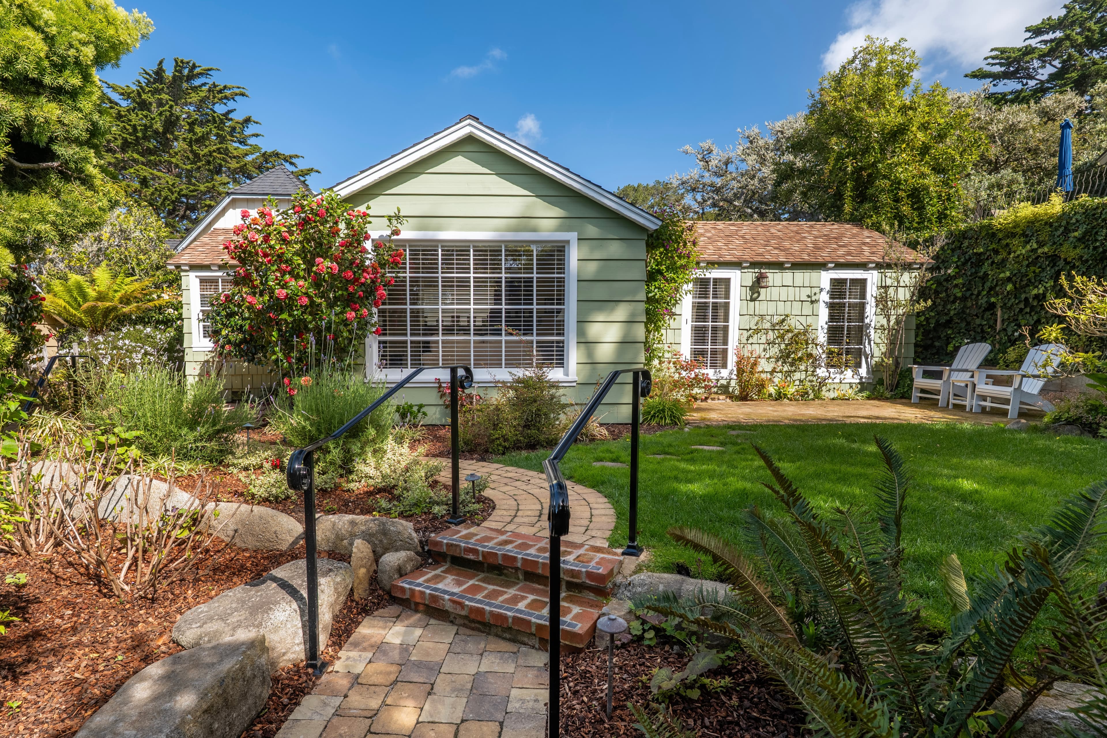 Exterior view of a charming pale-green cottage with a gabled roof and white-trimmed windows, featuring a paver path leading up brick steps through a lush green lawn and garden.