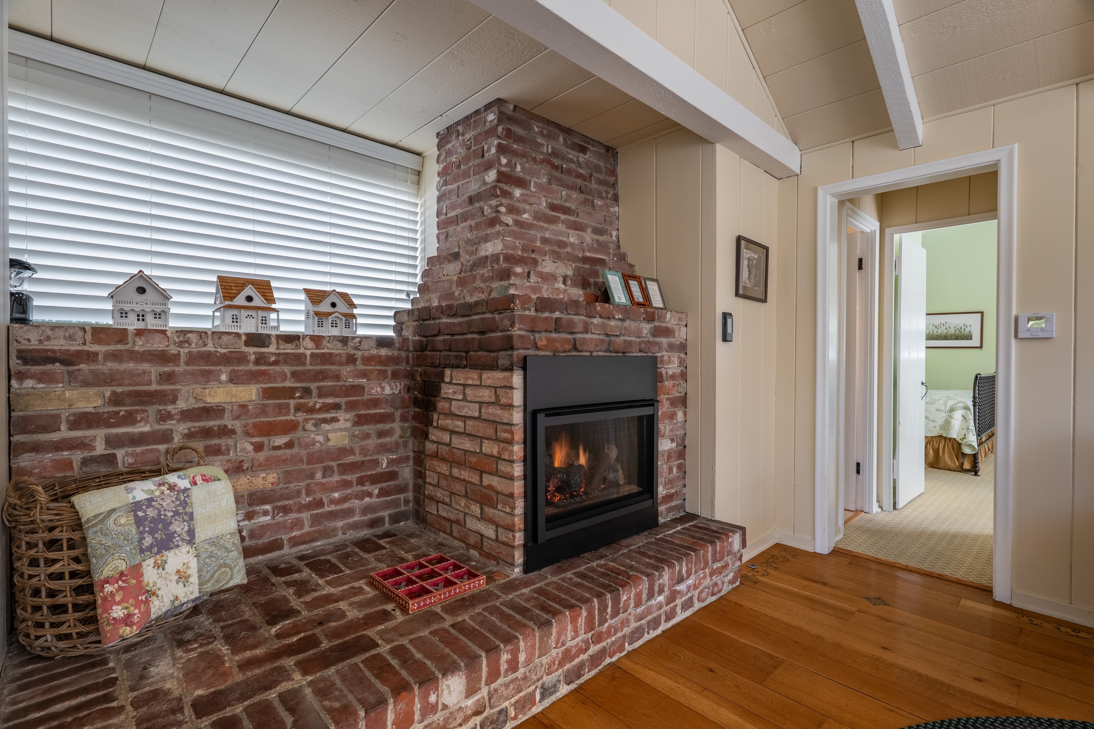A cozy living room with a vaulted white-beamed ceiling and hardwood floors, featuring a large brick fireplace with a black insert, and a partial view into the bedroom.