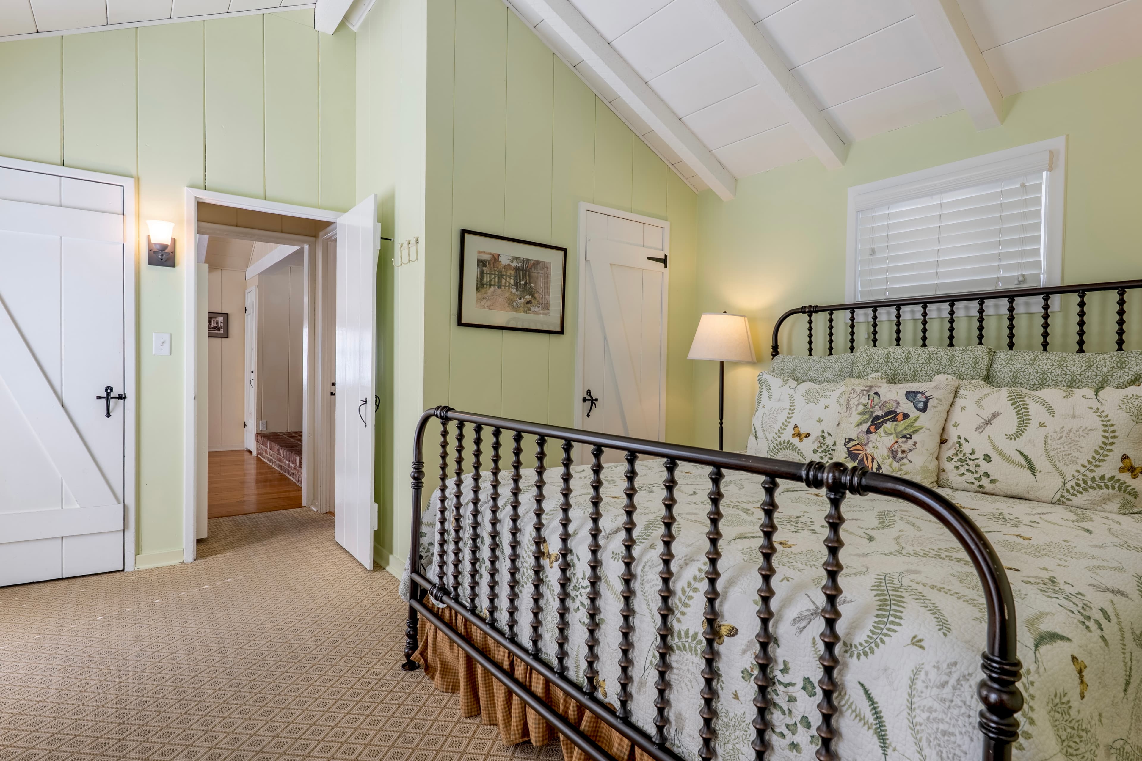 A bright bedroom with pale green walls and a vaulted white-beamed ceiling, featuring a dark metal bed frame with a patterned quilt and a view through an open door into a hallway.