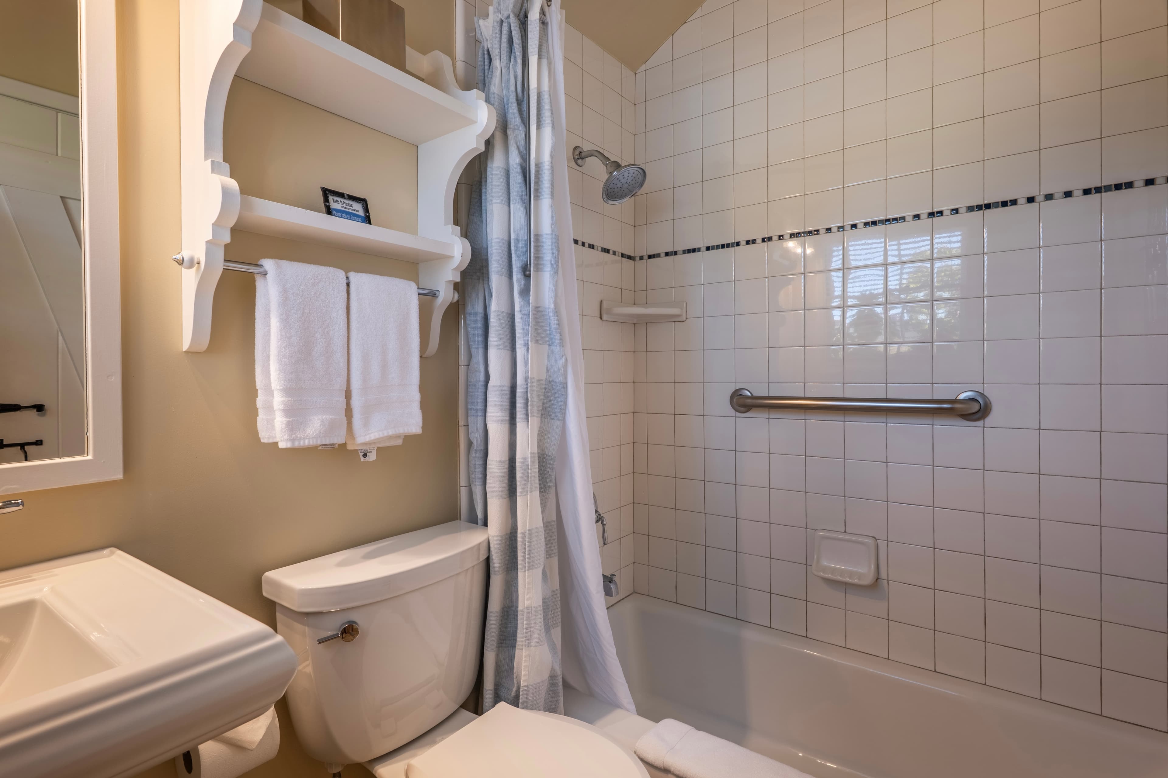 A classic cottage bathroom featuring a white tiled bathtub and shower with a safety grab bar, a white pedestal sink, and a vintage-style scalloped wall shelf holding fresh white towels.