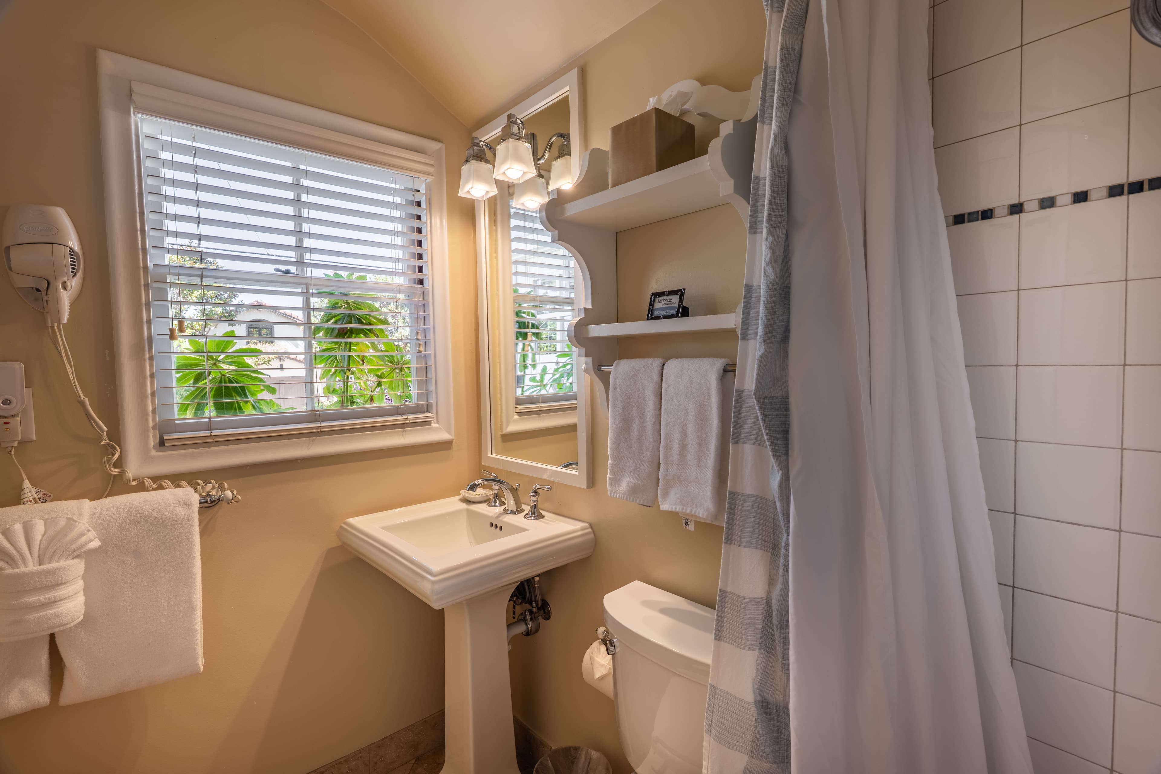 A classic cottage bathroom featuring a pedestal sink, a wall-mounted hairdryer, and a window with white blinds overlooking greenery. A white scalloped shelf holds fresh towels next to a shower with a blue and white checkered curtain.