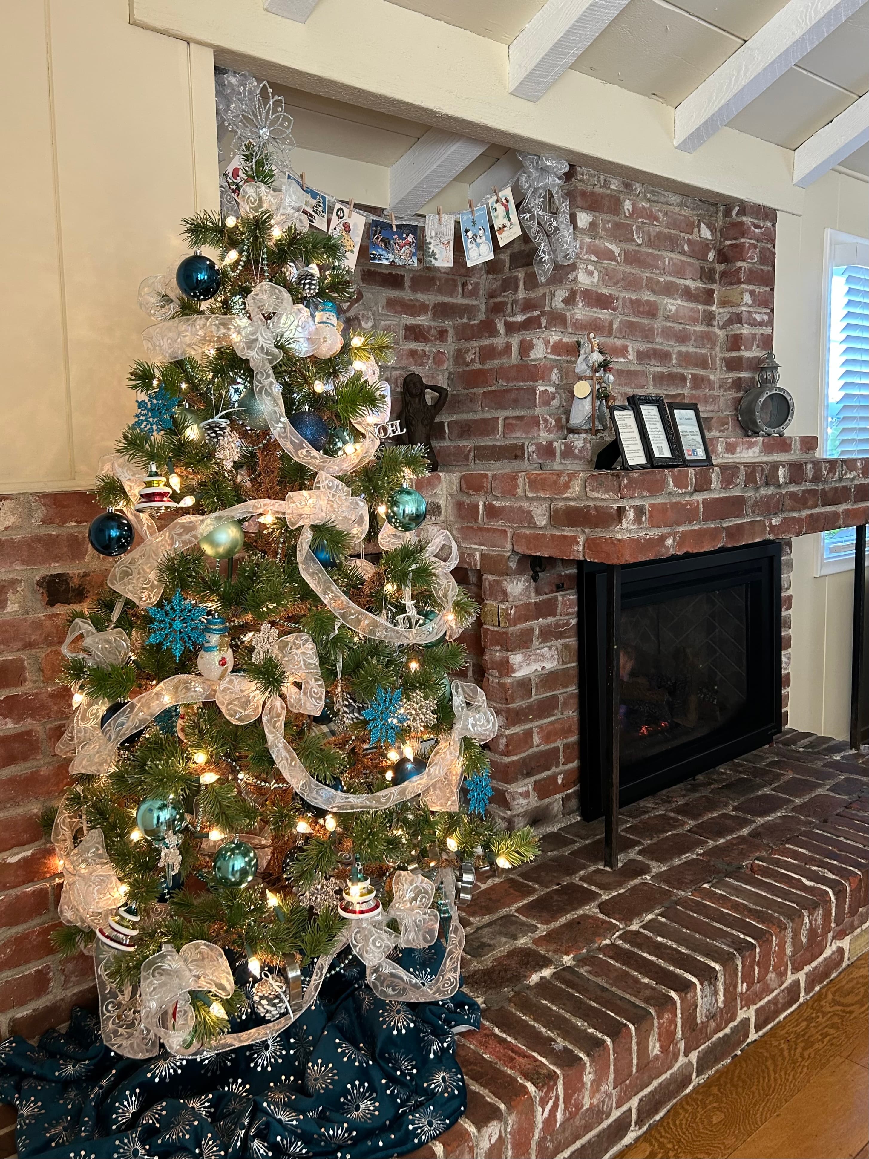 For our December guests, a festive Christmas tree decorated with blue and silver ornaments and white ribbon, standing in front of a large brick fireplace with a black insert and hearth.