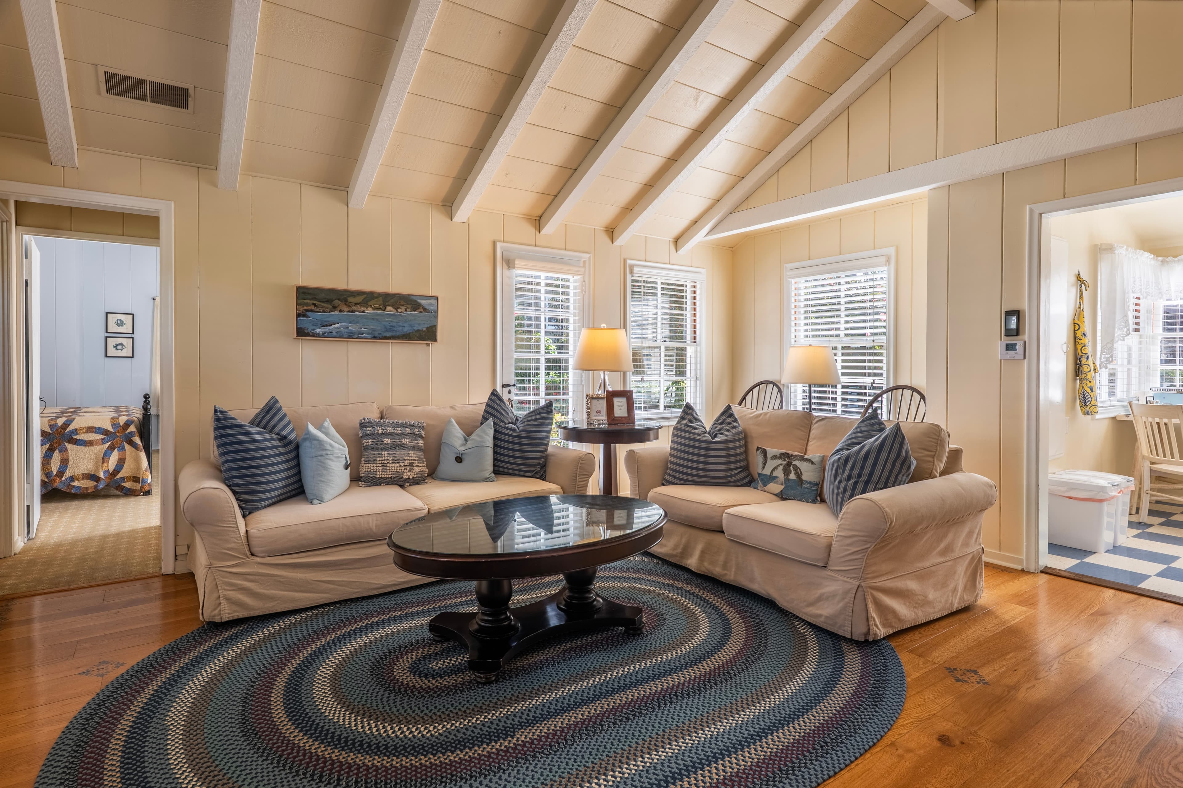 A cozy living room with beige couches, blue throw pillows, white beamed ceiling and a view into the kitchen and the bedroom.