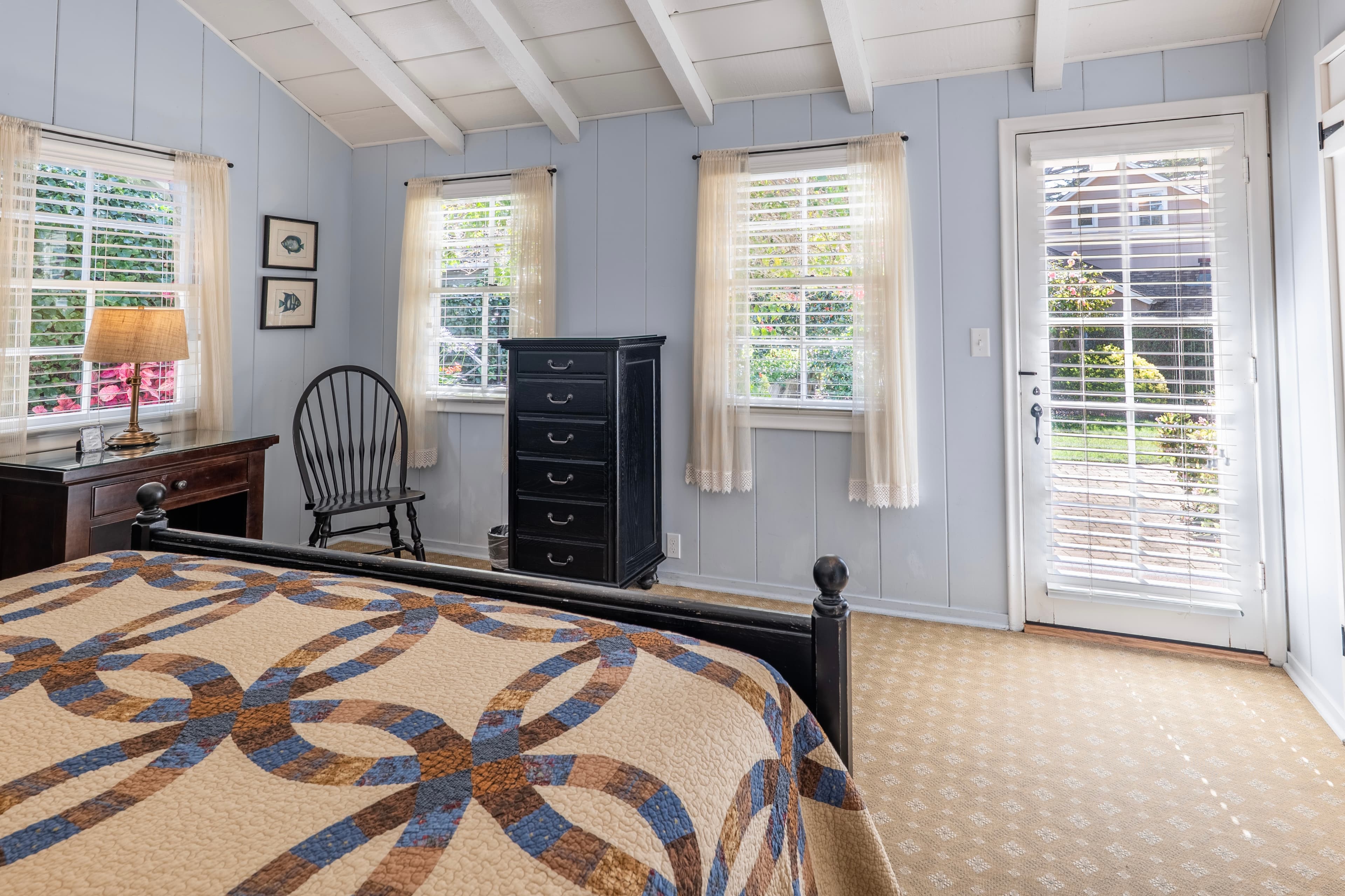 A bright bedroom with light blue wood-paneled walls and a white vaulted beamed ceiling, featuring three windows with sheer curtains and a glass-paneled door leading outside.