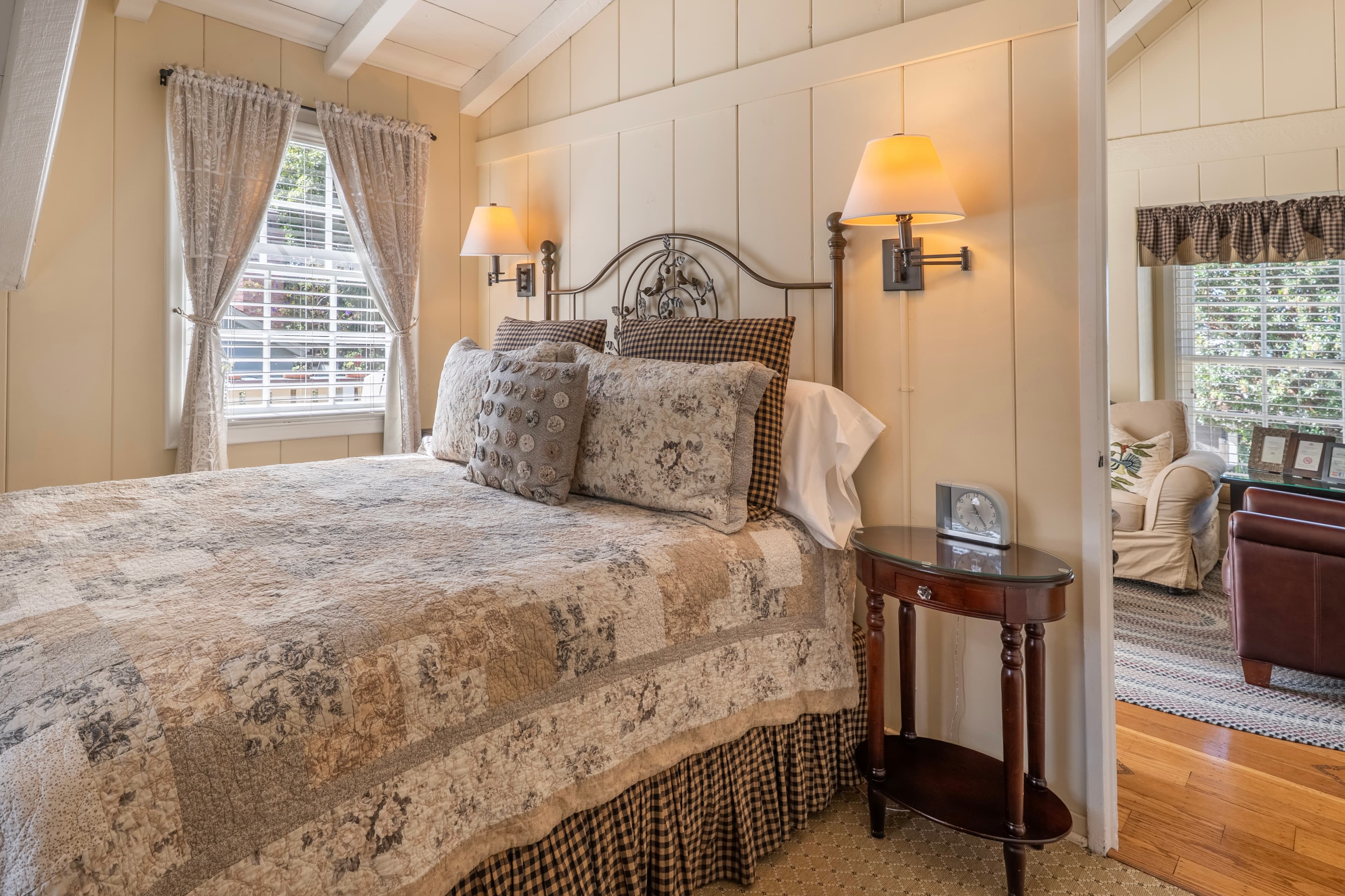 A cozy guest bedroom with sloped cream walls and vaulted white-beamed ceilings, featuring a bed with a patterned quilt, an ornate metal headboard, and bedside lamps.