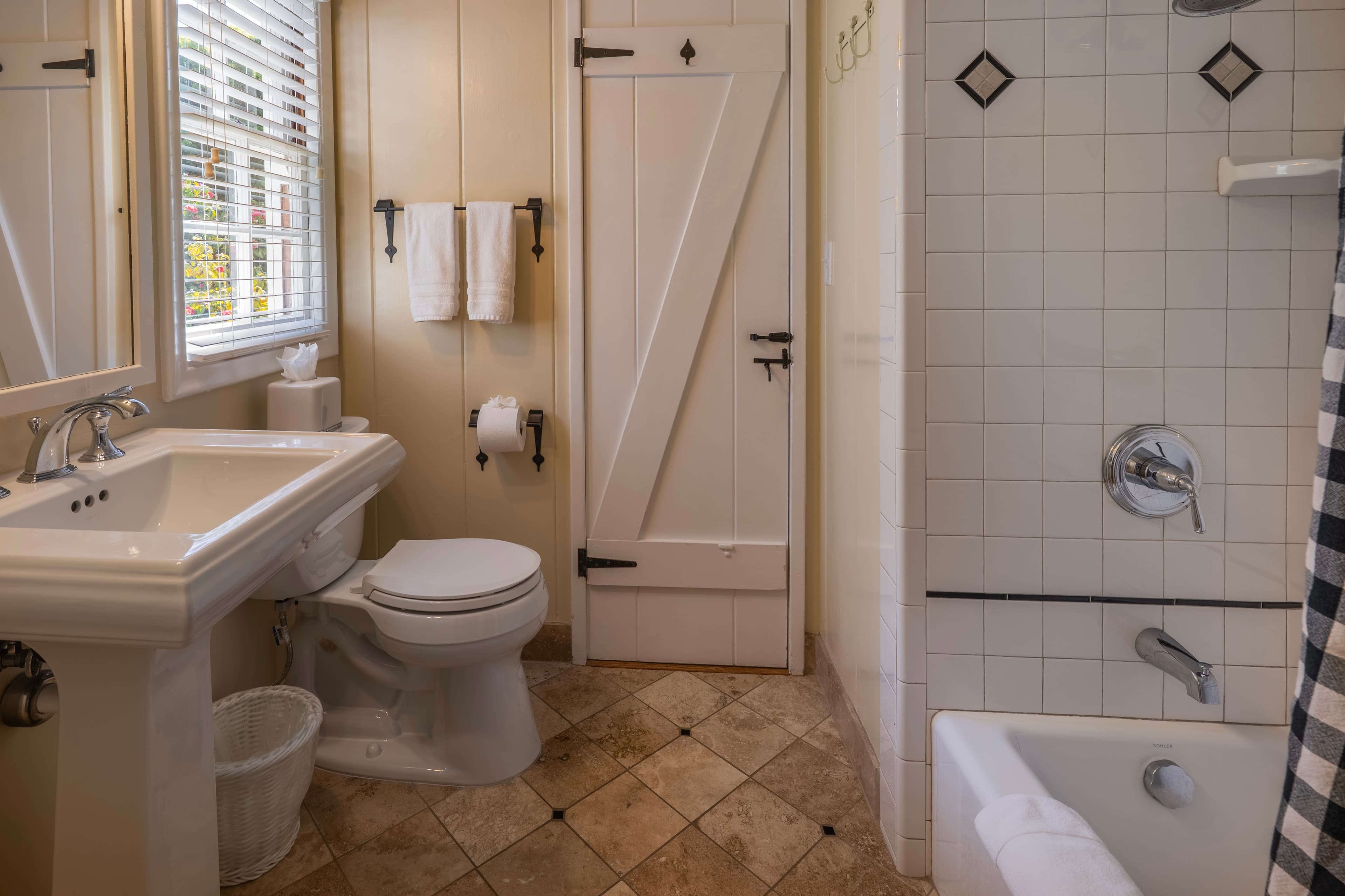 A classic bathroom with tan walls and large tan floor tiles, featuring a white pedestal sink, a toilet, and a white tiled shower with decorative black diamond accents and a black and white checkered curtain.
