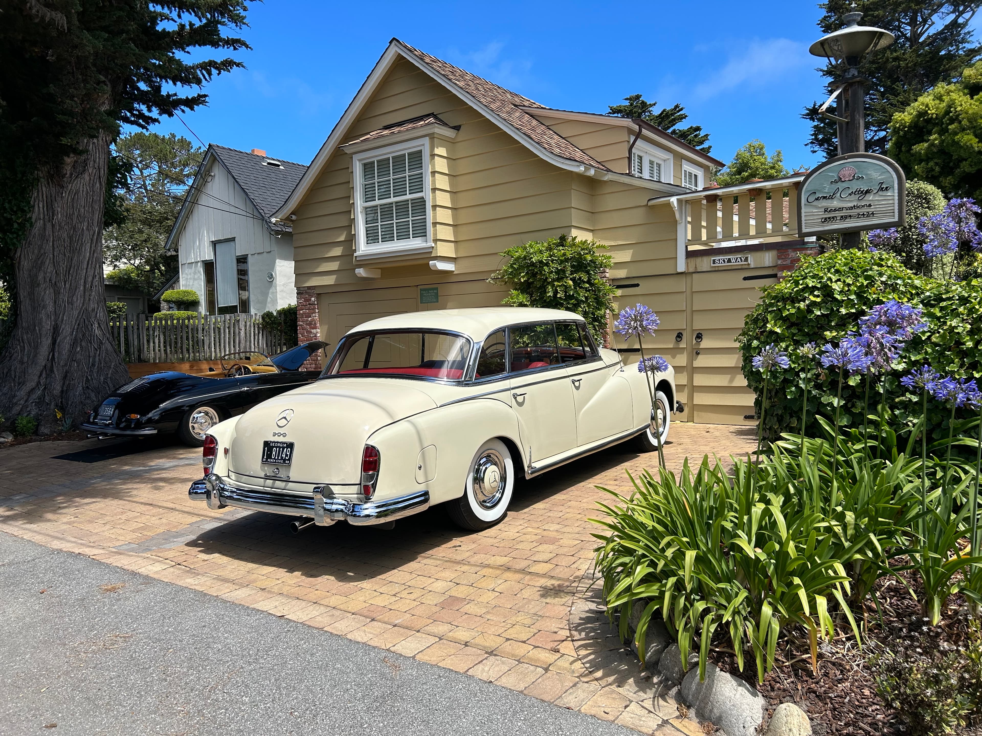 Exterior view of a tan two-story cottage with a wooden balcony and flower boxes, with a vintage cream-colored car parked on the stone driveway in front of the building.