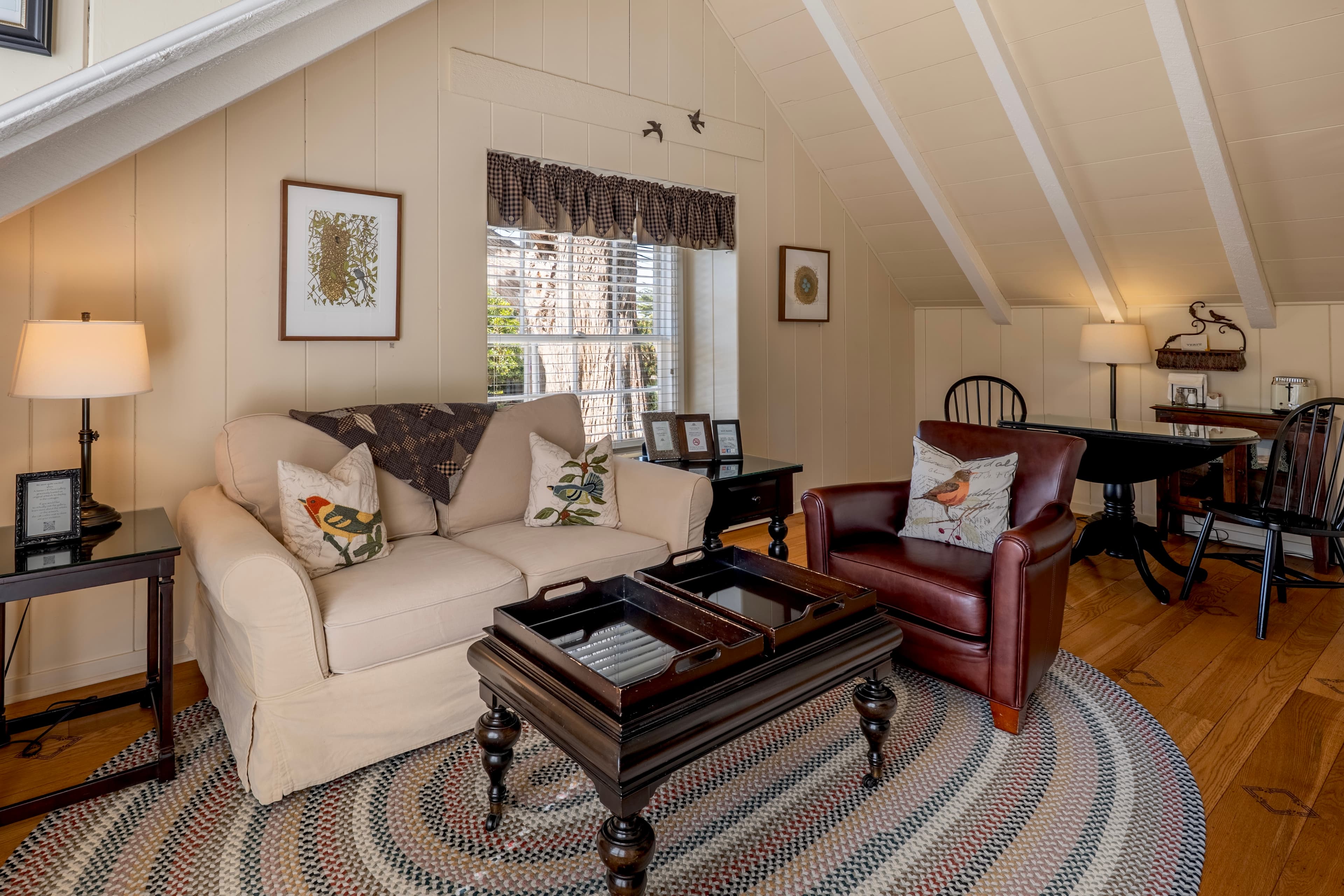 A cozy upper-level sitting area with vaulted white-beamed ceilings, featuring a neutral loveseat, a leather armchair, and a dark wood coffee table with tray tops on a circular braided rug.