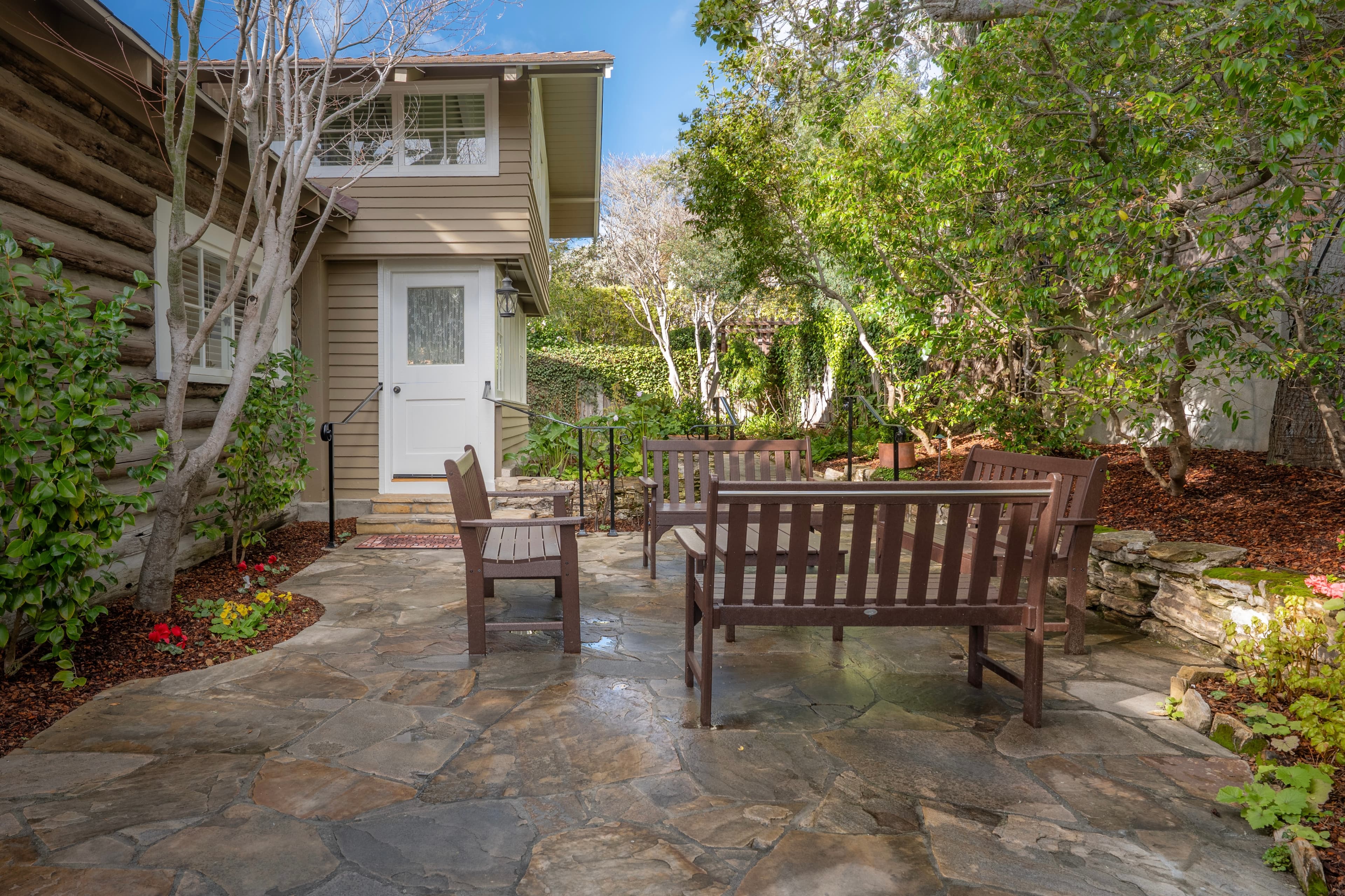 A stone patio with wooden benches surrounded by greenery and a house in the background.