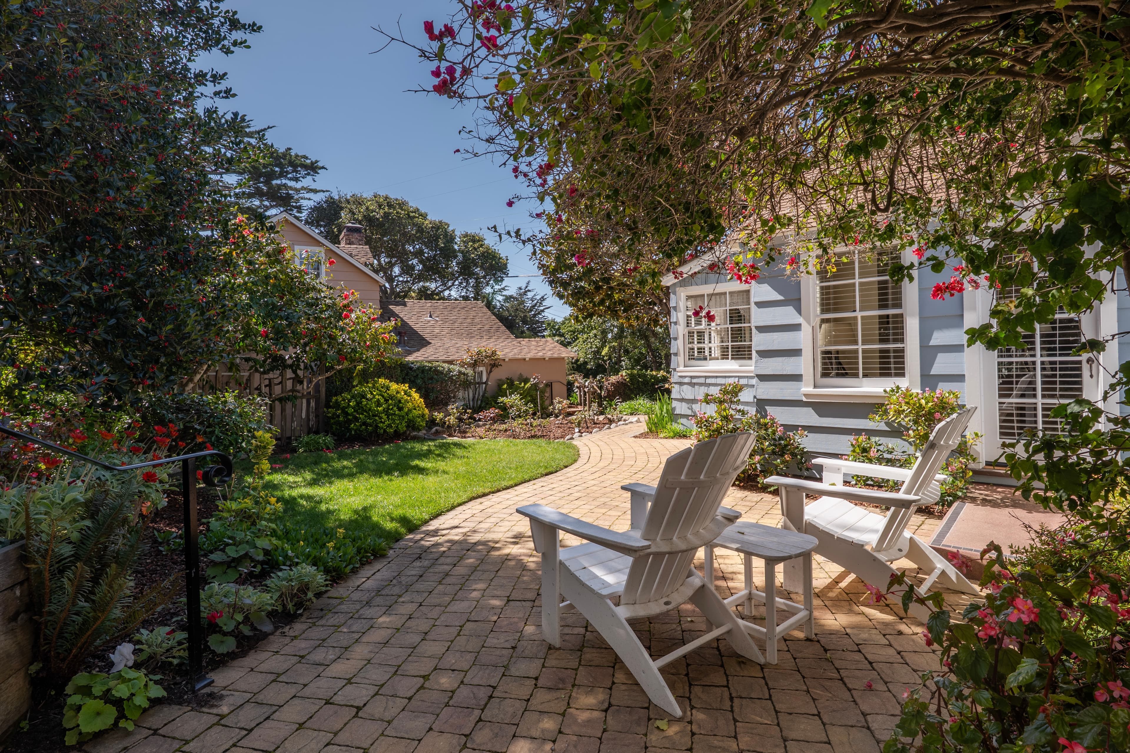 Two white Adirondack chairs and a small side table on a paver patio, surrounded by a lush green lawn and garden with a blue cottage in the background.