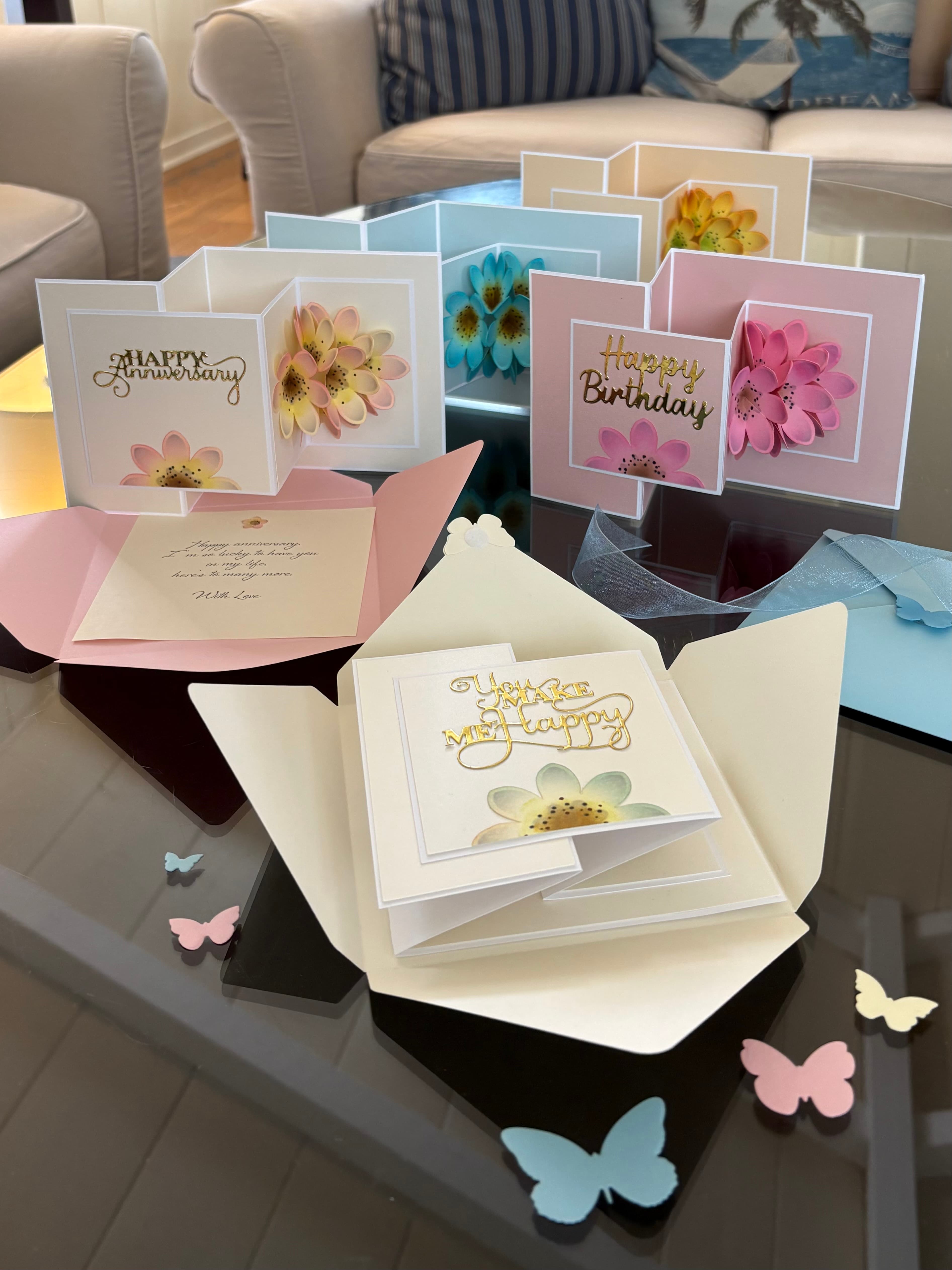 A collection of colorful pop-up greeting cards for birthdays and anniversaries displayed on a glass table, surrounded by small decorative butterflies.
