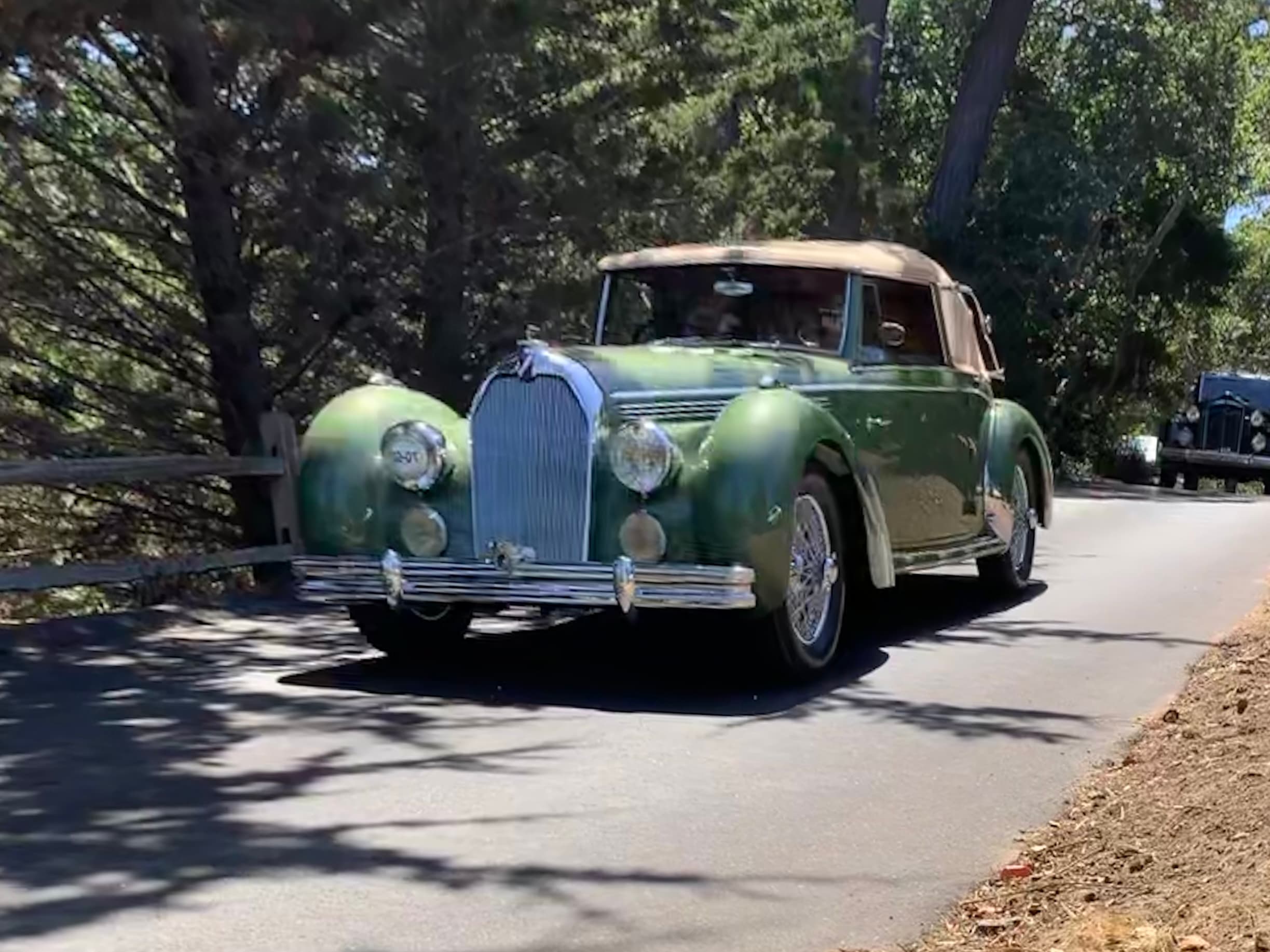 An antique green convertible car drives along a tree-lined road.