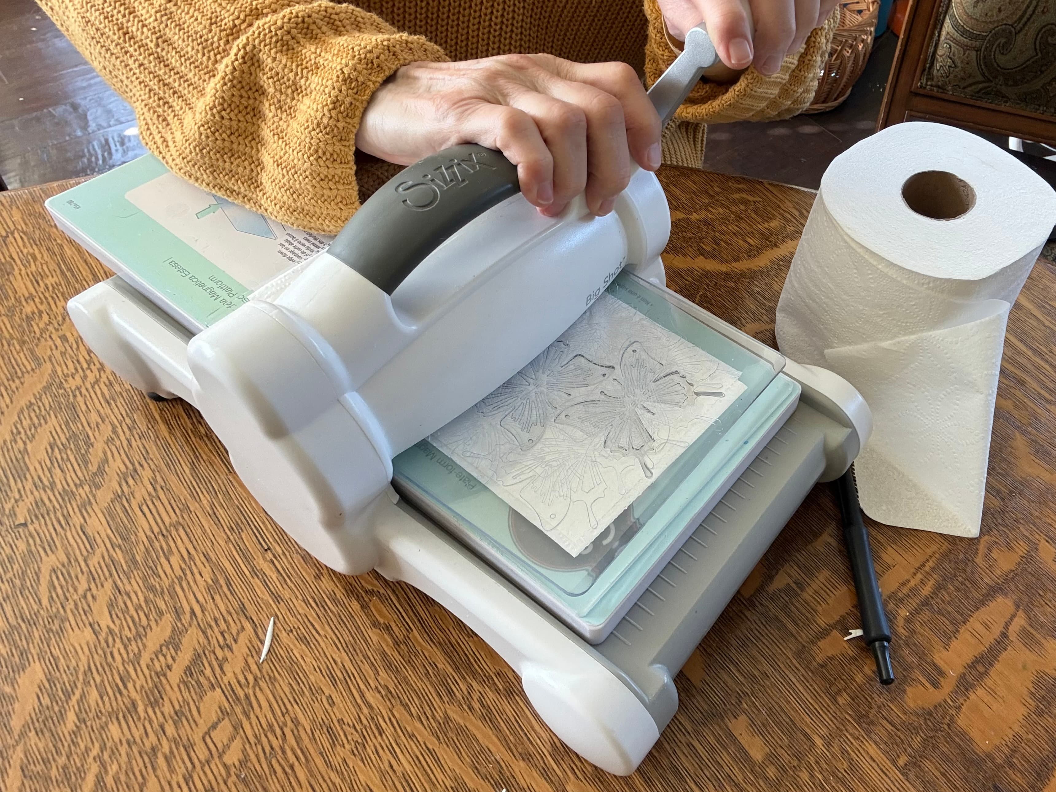 A person is using a die-cutting machine on a wooden table with paper towels nearby.