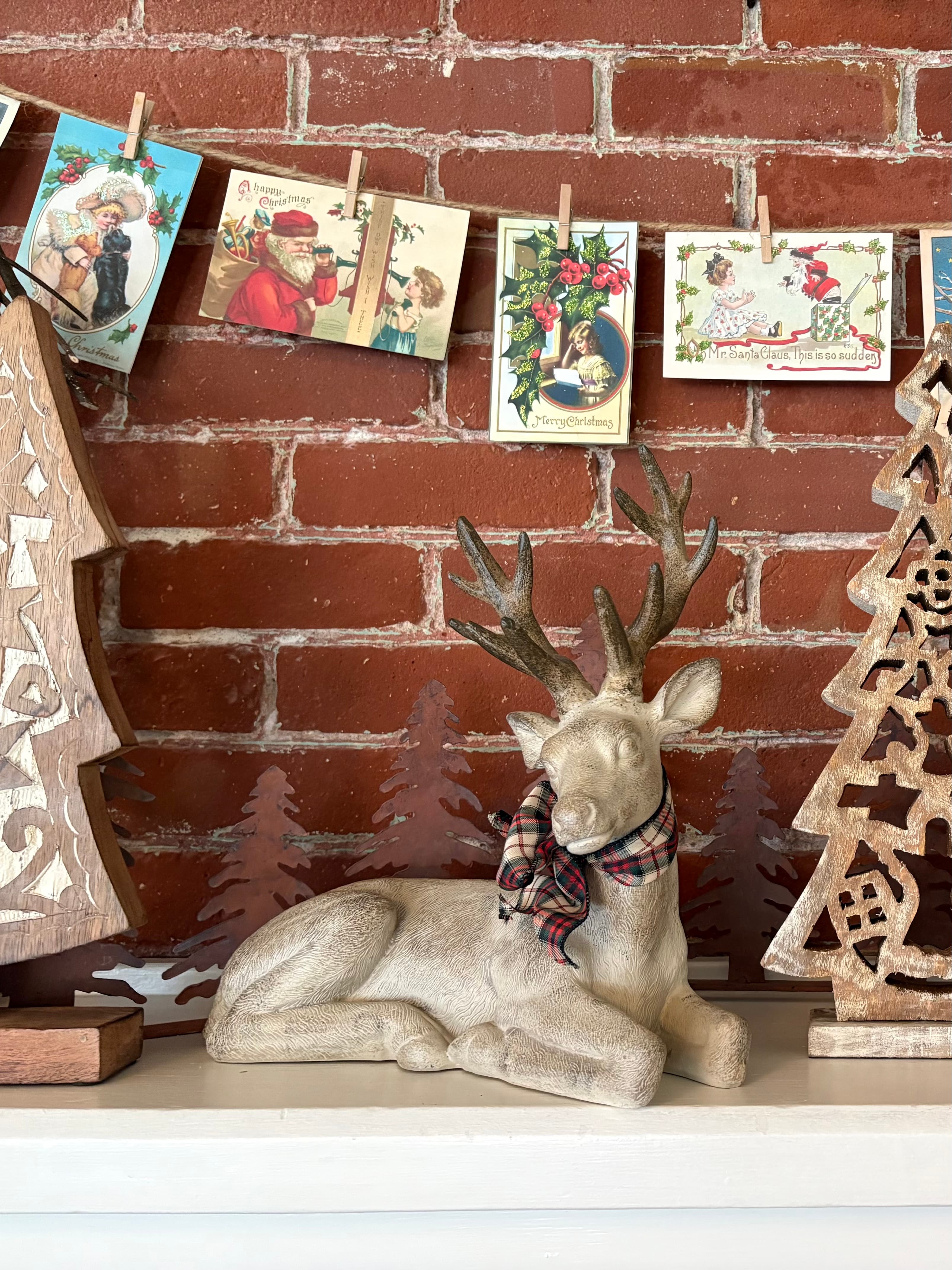 A decorative reindeer statue wearing a plaid scarf is positioned among wooden Christmas trees and handmade "vintage" holiday cards against a brick wall.
