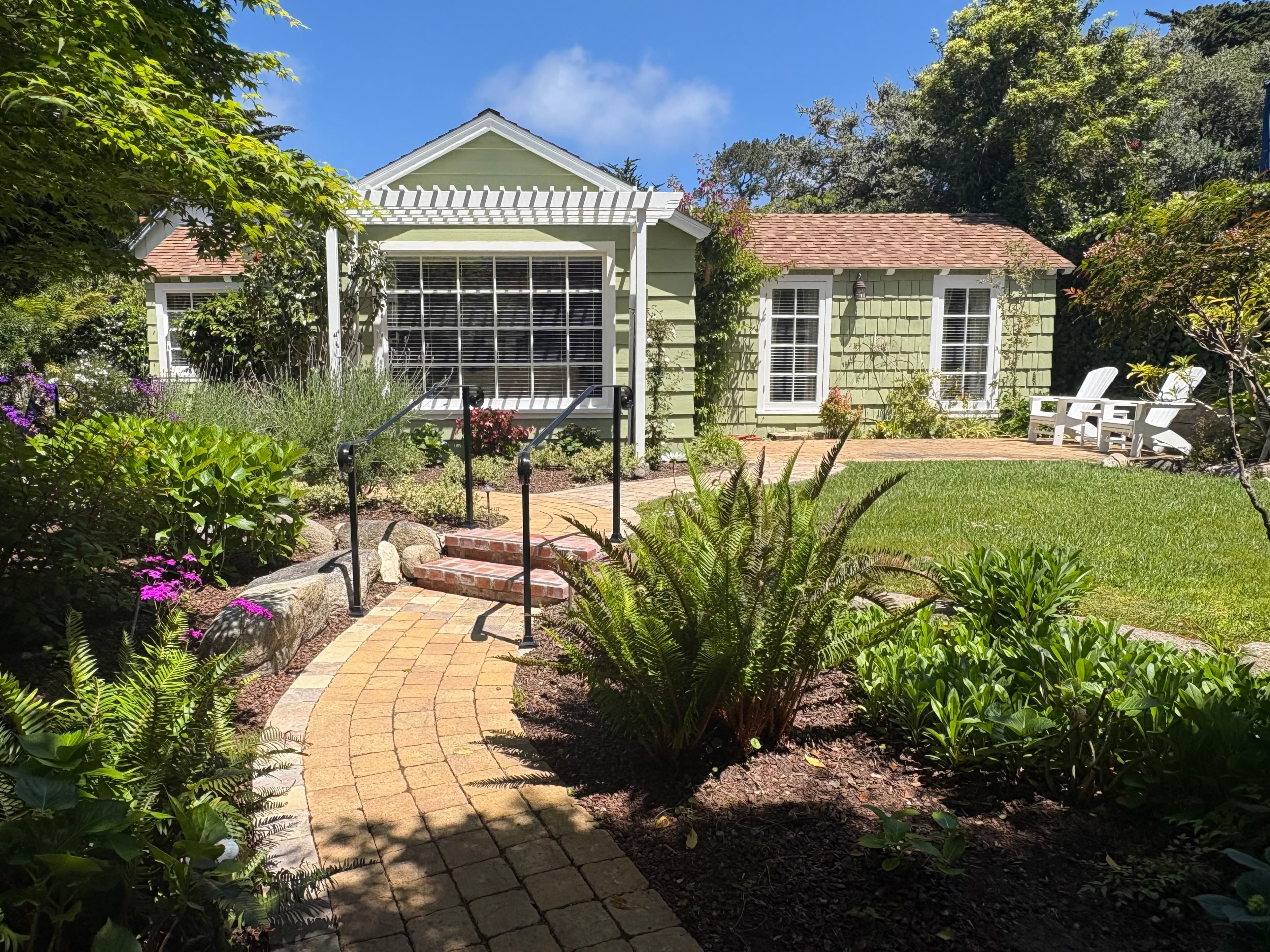 Exterior view of a charming pale-green cottage with a gabled roof and white-trimmed windows, featuring a paver path leading up brick steps through a lush green lawn and garden.