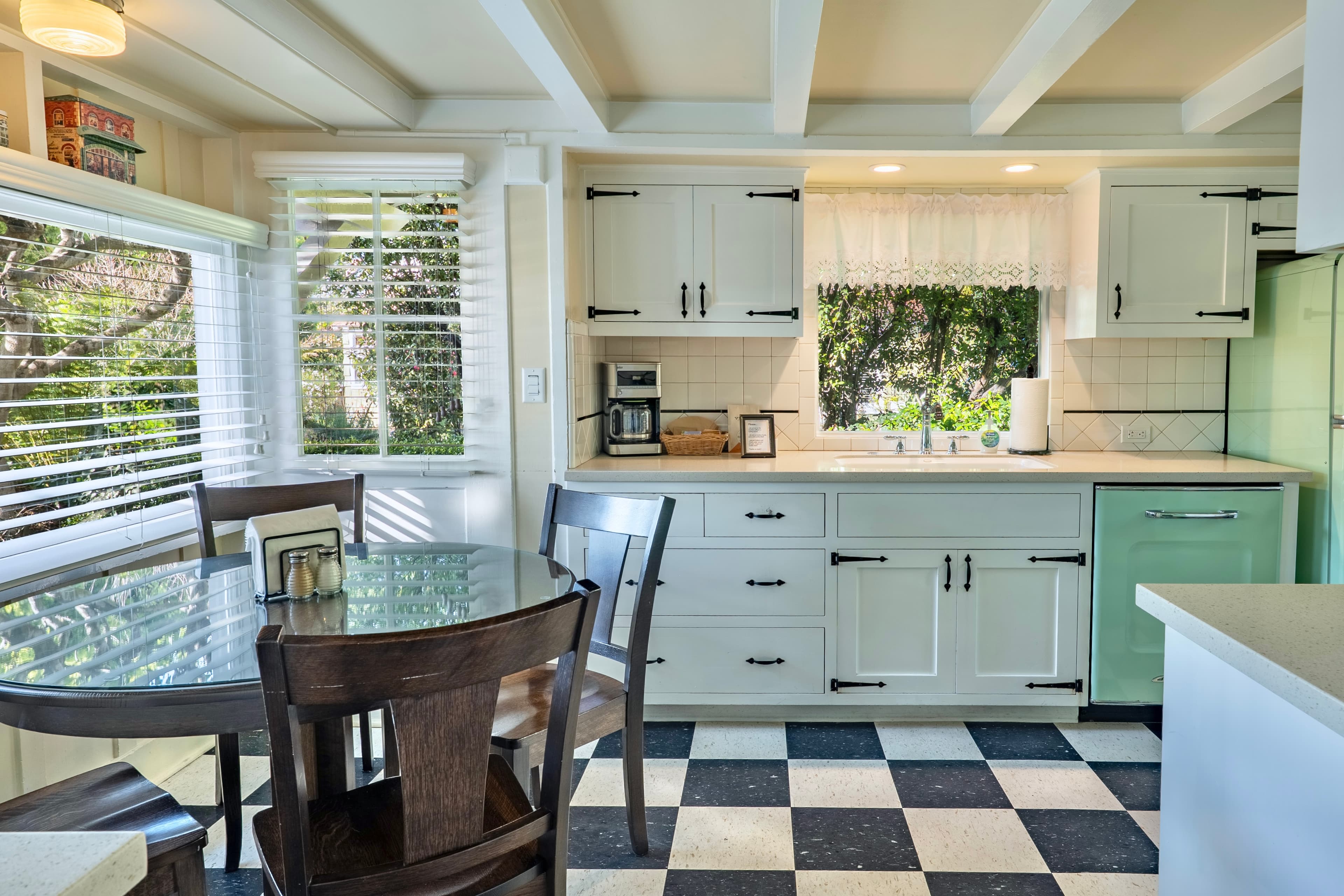 A bright kitchen features a checkered floor, wooden dining table, and green appliances with views of greenery through the windows.