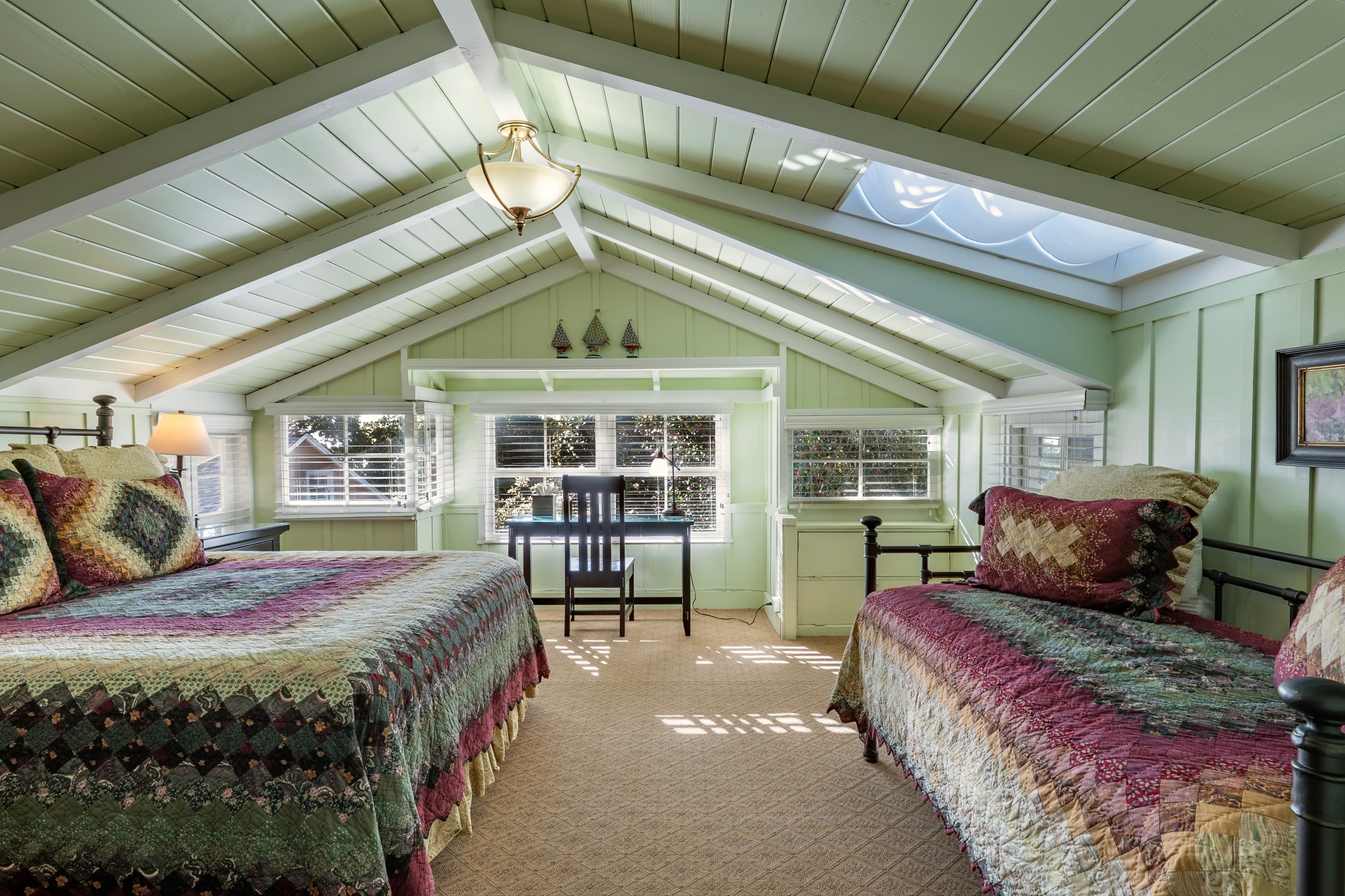 A cozy attic bedroom featuring two beds with colorful quilts and a desk beside large windows.