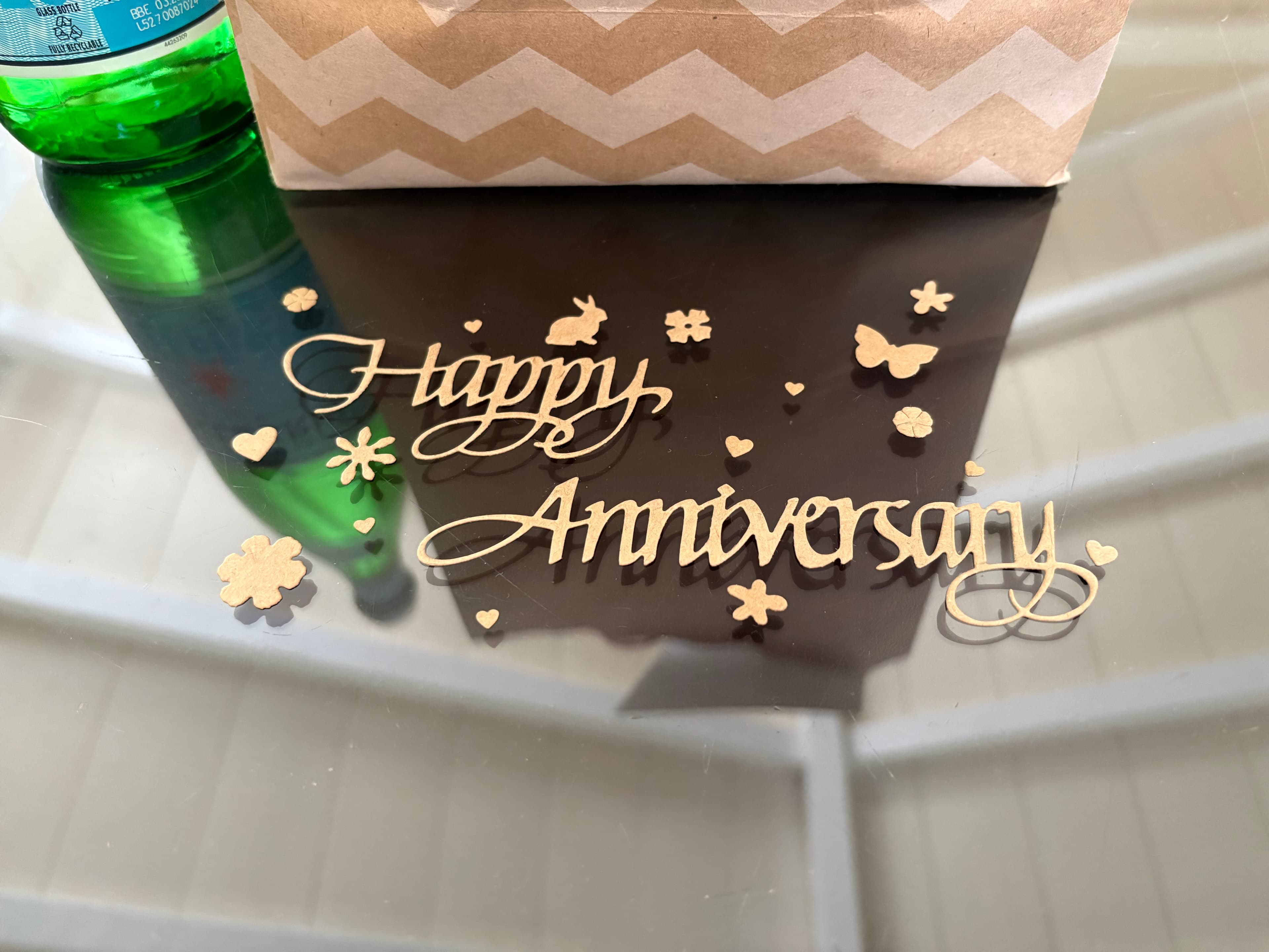 A reflective surface displays a decorative "Happy Anniversary" sign surrounded by small hearts and flowers, with a green bottle and a kraft paper bag nearby.